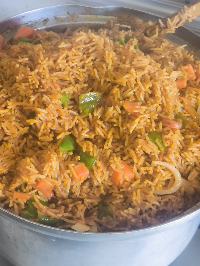 The image displays a close-up view of a large, metallic cooking pot filled with Jollof rice. The rice is a reddish-orange color, mixed with distinct pieces of diced orange carrots, chopped green bell peppers, and translucent white onion rings. Individual long-grain rice kernels are visible throughout the dish. A wooden utensil, possibly a spoon or spatula, is partially submerged in the rice on the upper right side of the pot. The pot's metallic rim is visible at the top and its curved side at the bottom of the frame. The background is a blurred, light-toned surface, indicative of an indoor setting, likely a kitchen. No individuals or active interactions are depicted. The specific location within Nicosia, Northern Cyprus / Cyprus, cannot be determined from the visual information provided.