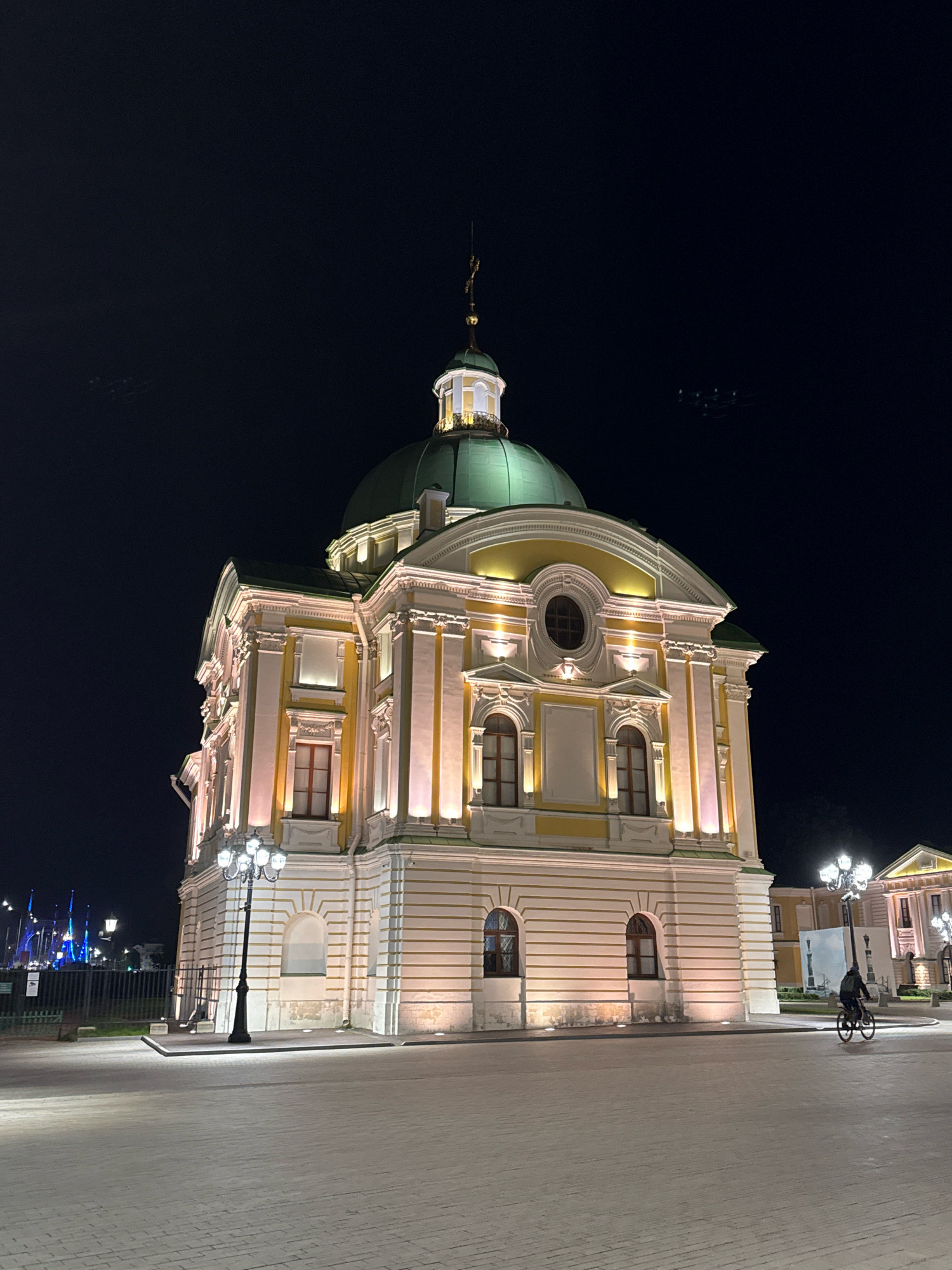 The image captures a night scene in the city of Tver, Russia, featuring the Bartholomew Cathedral. The cathedral is illuminated by warm lights, highlighting its ornate architecture, including a prominent green dome and yellow accents. In the foreground, a cobblestone plaza is visible, with a vintage-style lamppost standing to the left of the cathedral. To the right of the cathedral, a lone individual is seen riding a bicycle on the plaza. In the background, more illuminated buildings and structures, possibly part of the same architectural complex or nearby structures, are visible. The overall atmosphere is serene and illuminated, suggesting a peaceful evening in the city.