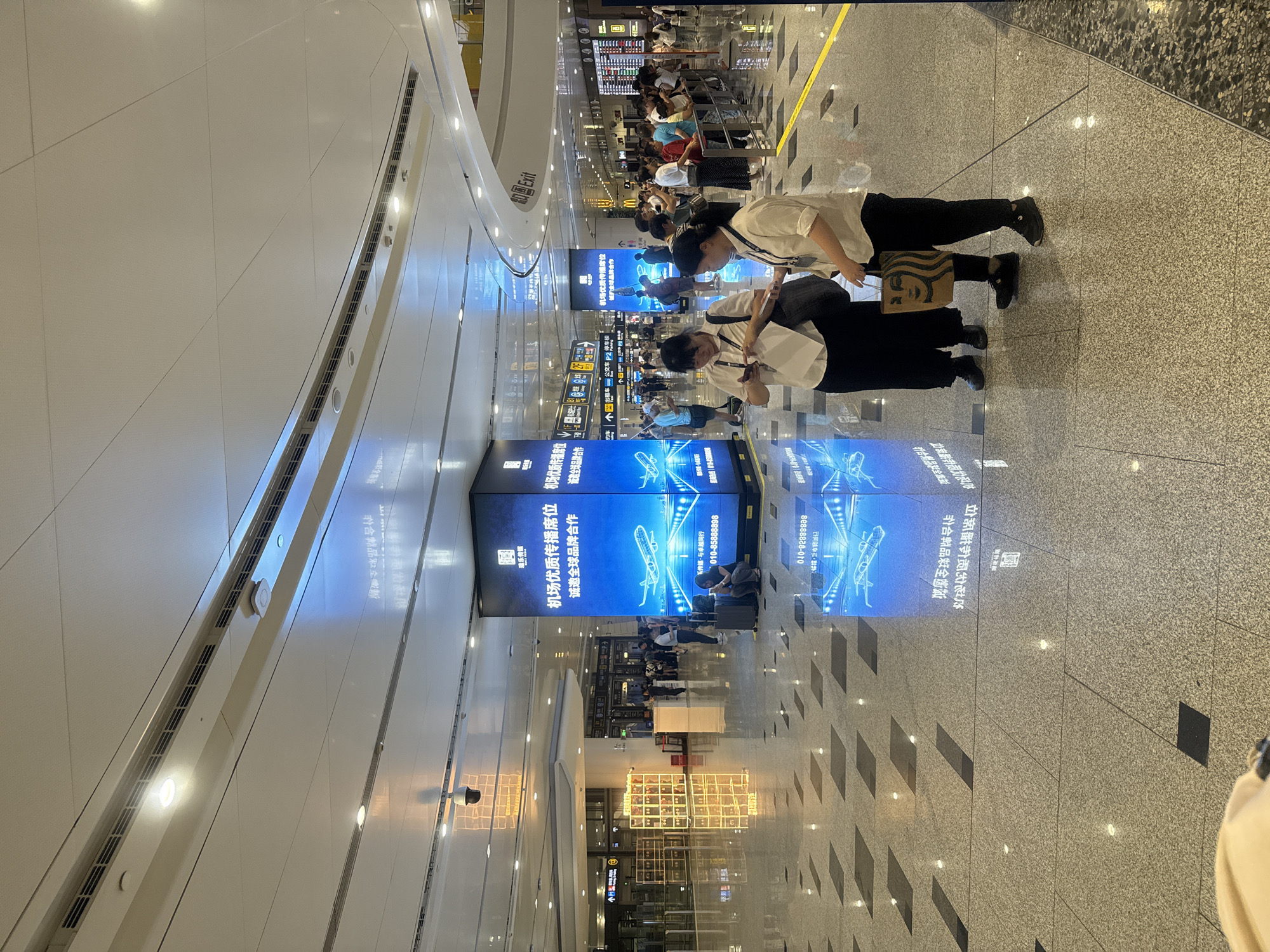 The image captures the interior of a modern airport terminal, likely in Guangyang District, China, characterized by its polished granite floor tiles with a speckled grey and black pattern, reflecting the bright overhead lighting and large digital displays. In the foreground, two women are standing, facing slightly away from the camera. The woman on the left, wearing a white blouse and black pants, holds a white paper in her left hand and a dark bag on her shoulder. The woman on the right, also in a white blouse and black pants, is holding a brown paper shopping bag with a smiley face logo, identified as a Starbucks bag, in her right hand.
Further into the terminal, a large group of people are visible, some standing and others walking. They are dispersed throughout the space, with some gathered near information desks or waiting areas. A prominent, illuminated blue digital billboard stands tall in the middle ground, displaying images of airplanes and Chinese text. Another similar billboard is partially visible to its left. In the background, there are various signs and displays, including what appears to be a departure board with flight information and signage pointing towards an "Exit." Security cameras are mounted on the ceiling. The overall environment suggests a busy airport setting with a clean, contemporary design.