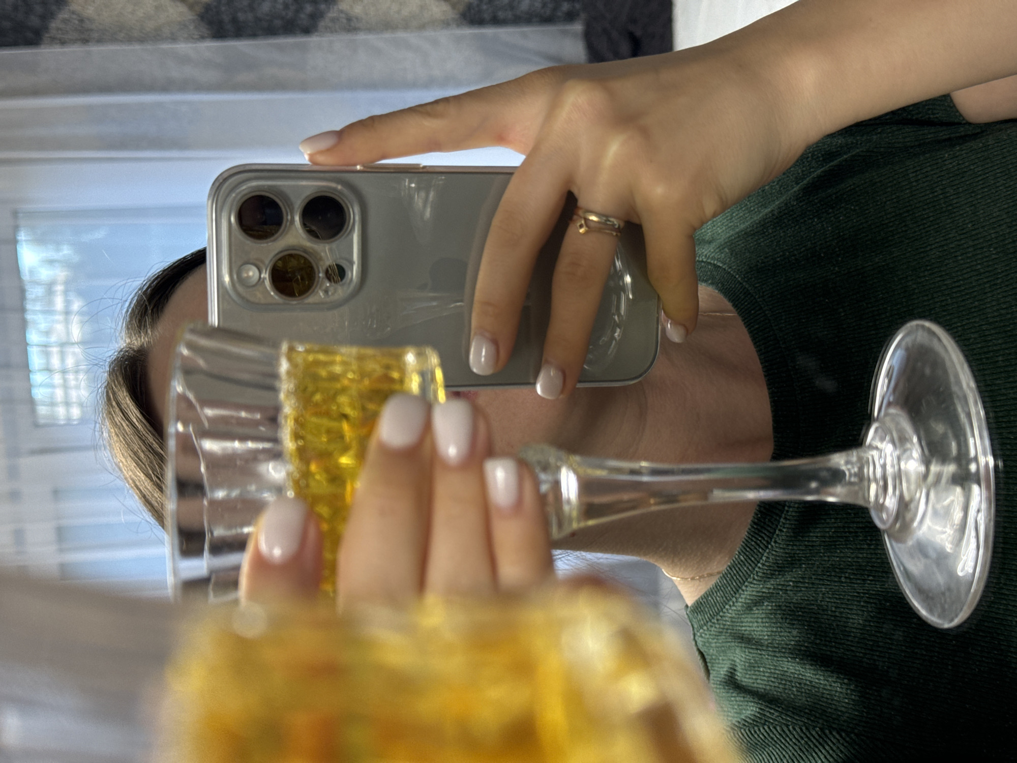 A woman is taking a selfie in a mirror. She is holding a glass of amber liquid in her left hand and a smartphone in her right hand. She is wearing a dark green ribbed t-shirt and a ring on her right ring finger. The background appears to be an indoor setting with sheer curtains and a patterned wall covering. The location is not identifiable as being in the city of Поназырево, Russia.