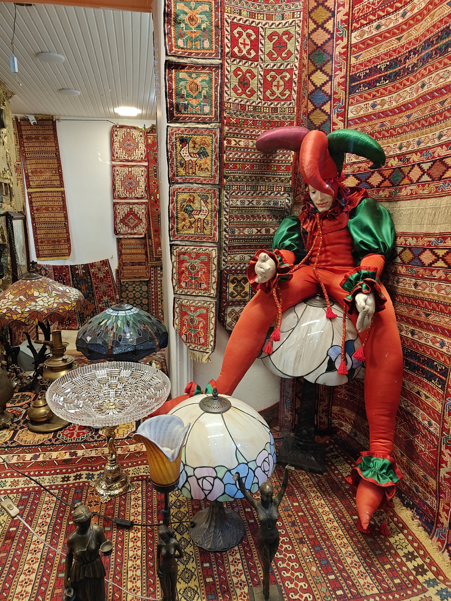 This media file captures an eclectic indoor shop display, likely in Tbilisi, Georgia, filled with ornate decorative items and textiles. The central subject is a life-sized mannequin, dressed as a jester, seated on a large, globe-shaped lamp. The jester wears a vibrant red and green costume with ruffled cuffs and a tri-pointed hat, its hands holding braided red cords with tassels.
The background and floor are extensively covered with numerous patterned rugs and tapestries in rich reds, greens, yellows, and blues, featuring geometric and stylized animal motifs typical of Caucasian or Middle Eastern designs. Many smaller, square rugs hang vertically, creating a textured wall.
In the foreground, several elaborate lamps are displayed. These include three Tiffany-style lamps with stained-glass shades: one with an autumn leaf pattern, another with a blue and green dragonfly design, and a third with a lighter floral pattern. A large, clear, cut-glass crystal bowl with intricate patterns is prominently placed on a decorative metal stand. Also visible are smaller bronze statues of female figures, one appearing to hold a bowl, scattered among the display items on the rug-covered floor.
The scene depicts a static retail or exhibition setup. The lighting appears artificial, suggesting an indoor setting or a shop window display at any time of day. The overall impression is one of a richly decorated space, showcasing a variety of handcrafted and antique-style goods. There is no visible text.