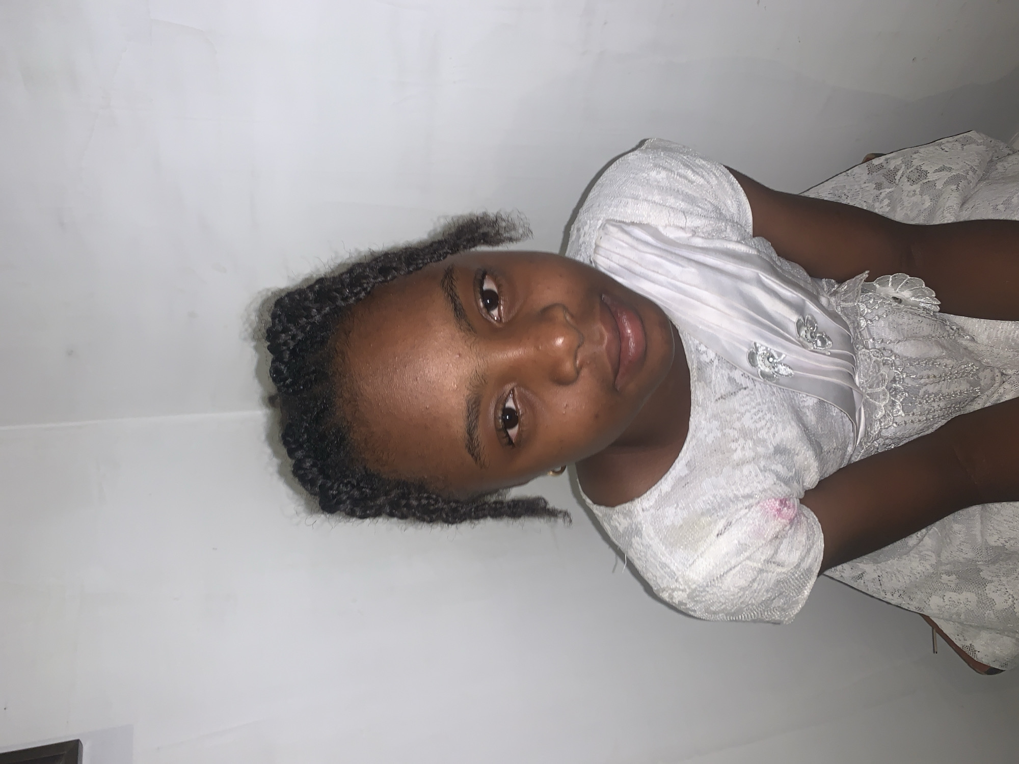 A young, dark-skinned girl with braided hair is indoors, wearing a white lace dress with satin accents. She is positioned against a white wall, and the photograph appears to have been taken in portrait mode. The location in Jos, Nigeria, is inferred and not directly discernible from the image.