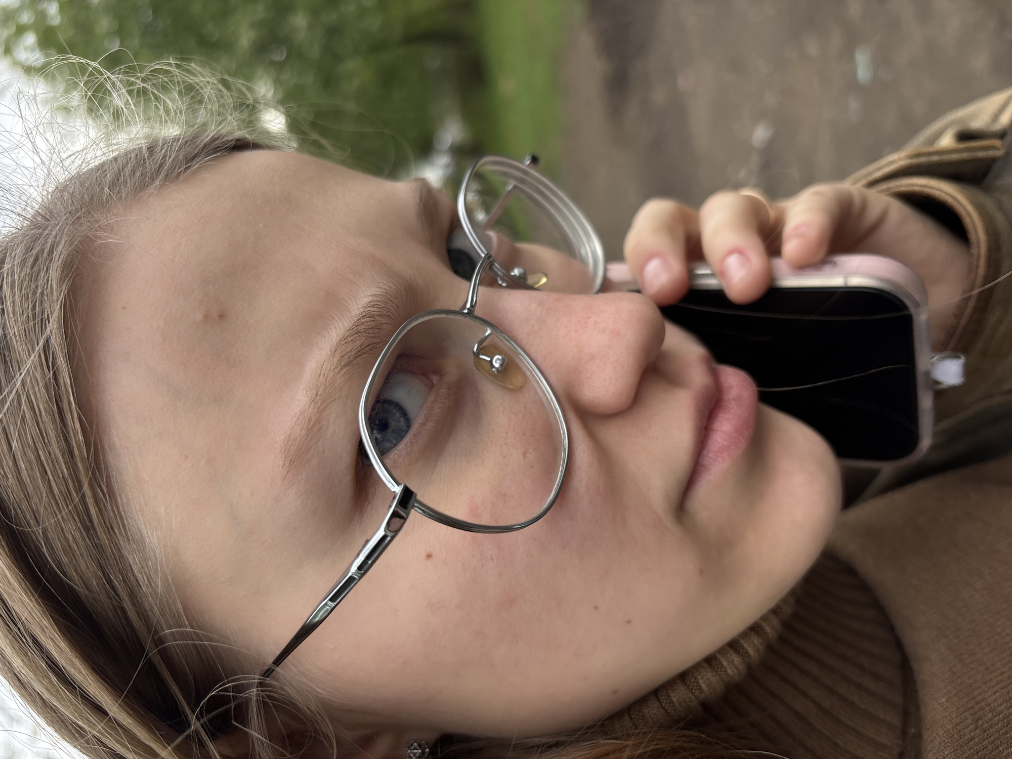 A close-up, angled shot depicts a young woman with fair skin and light blonde hair wearing glasses and holding a cell phone to her ear. She has blue eyes and a neutral expression. The glasses are silver-framed. Her turtleneck sweater is brown. The cell phone appears to be pink with a cracked screen. The background is slightly out of focus, showing greenery and indistinct brown surfaces. Without further evidence, determining a specific location in the city of Perm, Russia, is impossible.