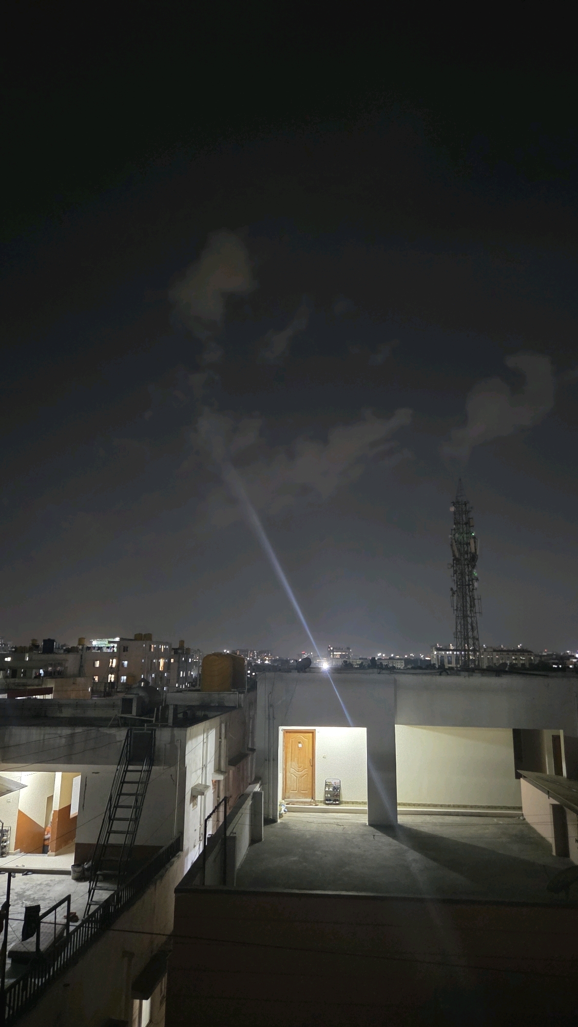 The image depicts a nighttime view of a cityscape, likely from a rooftop in Bengaluru, India. The sky is dark with scattered clouds illuminated by distant city lights and a strong beam of light cutting diagonally through the atmosphere. In the foreground, a concrete rooftop area is visible, with a building entrance featuring a wooden door and a small shoe rack. To the left, a metal staircase leads down to another level of the rooftop, with railings and some debris. Beyond the immediate rooftop, a row of buildings stretches across the horizon, with lights visible in their windows. Further in the distance, a tall telecommunications tower stands prominently, its structure illuminated. The overall scene conveys a sense of urban night life under a cloudy sky.