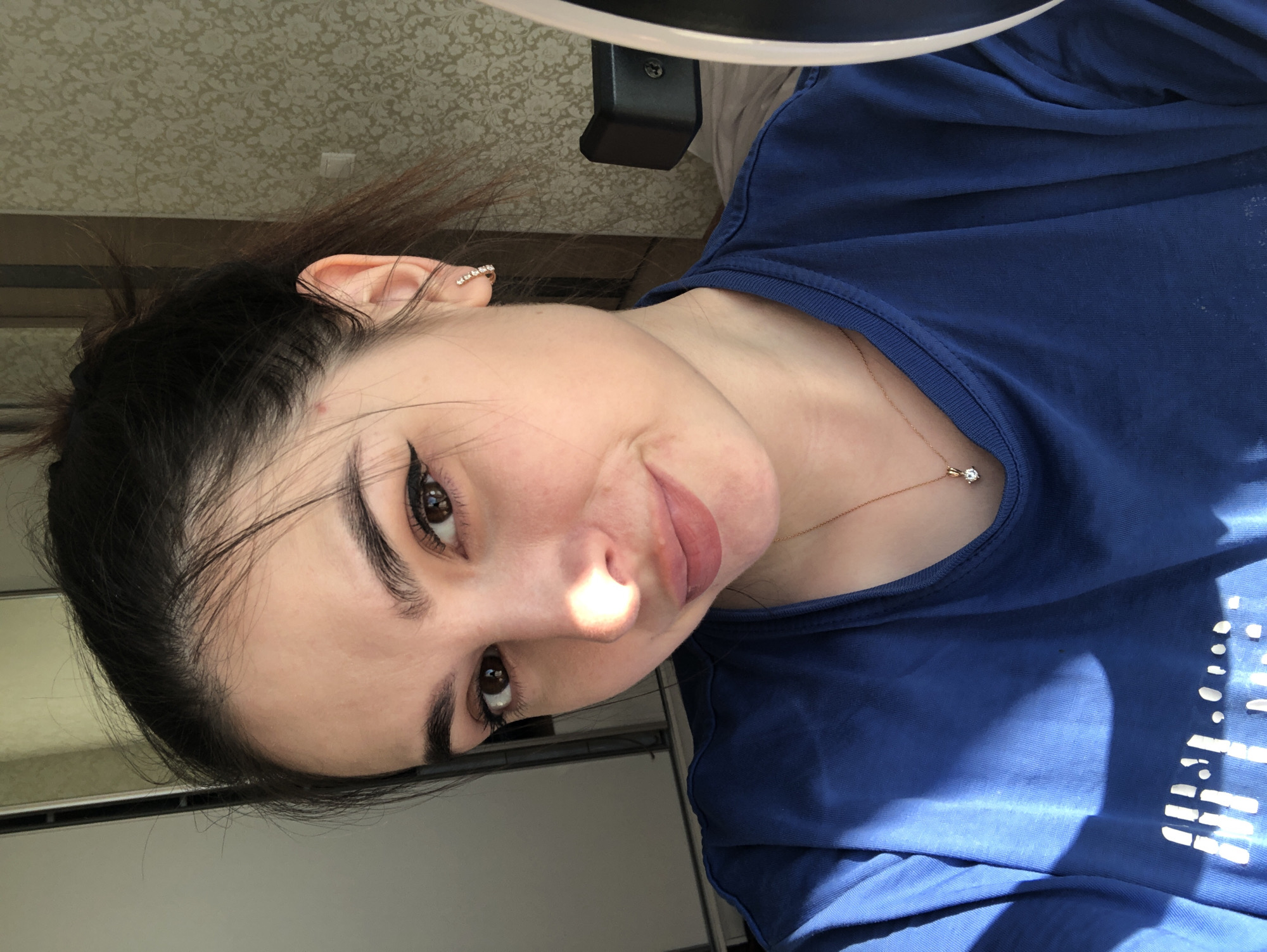 A young woman with fair skin, dark hair pulled back, and dark brown eyes is pictured in a close-up shot. She is wearing a blue t-shirt and a delicate gold necklace with a small diamond pendant. She has winged eyeliner and a light lip color. A sparkly earring is visible in her left ear. Bright light shines on her face, creating a highlight on her nose. The background is blurry but shows a patterned wallpaper and a light-colored wall. There is a black and white object partially visible above her head, possibly part of a lighting fixture or a piece of furniture. The photograph does not indicate a location in the city of Noviy, Russia.