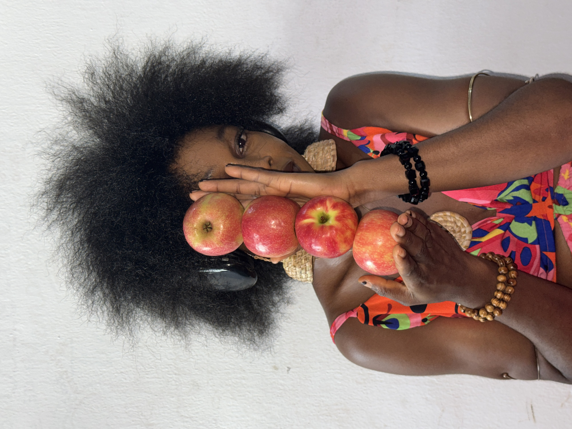The image is a portrait, originally oriented sideways, featuring a person with dark skin and voluminous, dark afro-textured hair against a plain, light-colored textured wall. The individual is positioned centrally in the frame.
**Subjects:** The primary subject is a person, likely a woman, with deep brown skin and a large, natural black afro. Their face is partially obscured by their hands, but one eye, gazing directly at the viewer, is visible. They are wearing a brightly colored garment with an abstract or floral pattern, featuring shades of orange, pink, blue, and green. Around their wrists and arms, they wear several accessories: a thin gold-toned bangle and a black beaded bracelet on one arm, and a wooden beaded bracelet on the other. Four fresh apples, predominantly red with hints of yellow, are held in their hands, forming a line across the lower part of the frame, in front of their chest and neck. A woven, natural-fiber element, possibly part of a necklace or accessory, is visible near their neck.
**Activity or event taking place:** The person is posing for a close-up portrait, holding the four apples prominently in their hands, which are positioned near their face. The pose suggests a deliberate artistic or fashion shoot.
**Notable details:** The lighting is soft and even, suggesting indoor conditions or an overcast day. The subject's striking afro hair is a prominent feature, creating a dynamic silhouette against the plain background. The vibrant colors of the apples and the person's clothing provide a contrast to the neutral wall. There are no discernible location cues within the image itself beyond the simple wall texture, though the provided context indicates Ado, Nigeria. No visible text is present. The expression in the visible eye is calm and direct.