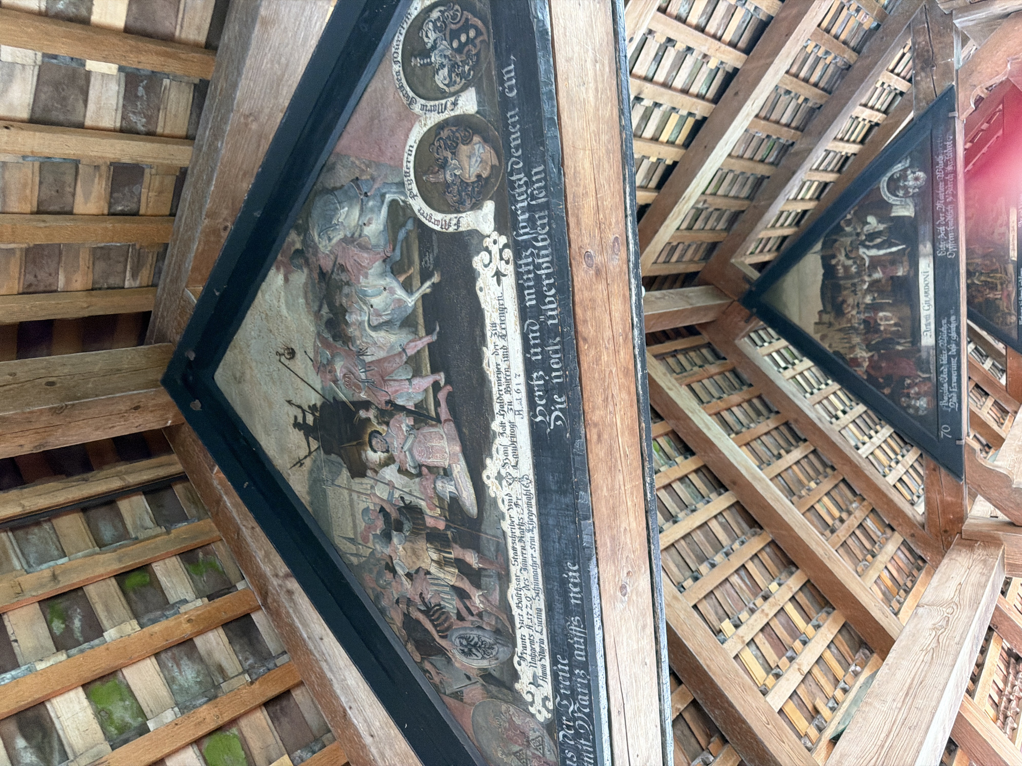The image shows the interior of a wooden roof structure, possibly a covered bridge. Two triangular panels with paintings are visible, angled towards each other. The upper panel depicts a historical scene with figures in armor, horses, and banners. Below the painting, there is text in a gothic script. The lower panel also features a painting of a historical scene with multiple figures, and text below it. The wooden roof structure itself is made of many small planks of wood, arranged in a lattice pattern. There are signs of wear and moss growth on some of the wooden elements. The overall impression is of an old, historic structure with preserved artwork. The location is identified as a covered bridge in an unknown city and country.