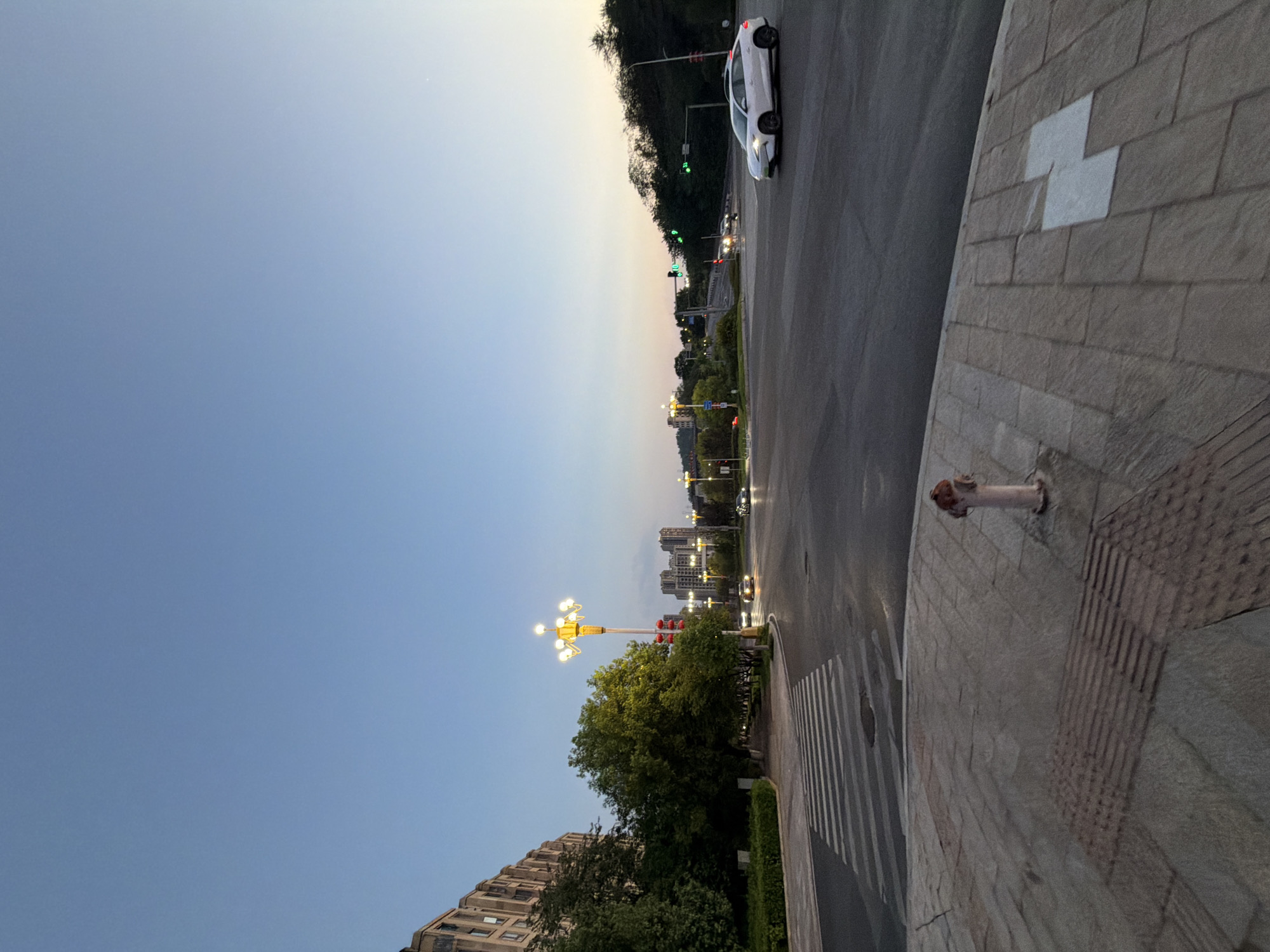 The image captures a street intersection in the city of Jiangjin, China, during dusk. The sky is a clear, pale blue transitioning to a warmer hue near the horizon, suggesting either sunrise or sunset. In the foreground, a paved pedestrian area with textured tactile paving and a single fire hydrant is visible. A crosswalk with white stripes stretches across the asphalt road.
Several vehicles are present on the road, including a white sedan on the right side of the intersection, appearing to be in motion, and other cars further in the distance, some with their headlights on. Traffic lights are visible, with some showing green signals.
On the left side of the image, a multi-story building with numerous windows stands adjacent to a dense line of green trees. A decorative street lamp with multiple illuminated bulbs is positioned near the pedestrian area. In the background, more buildings and trees line the horizon, with a hilly, tree-covered landscape visible on the far right. The overall scene depicts a quiet urban environment at twilight.
