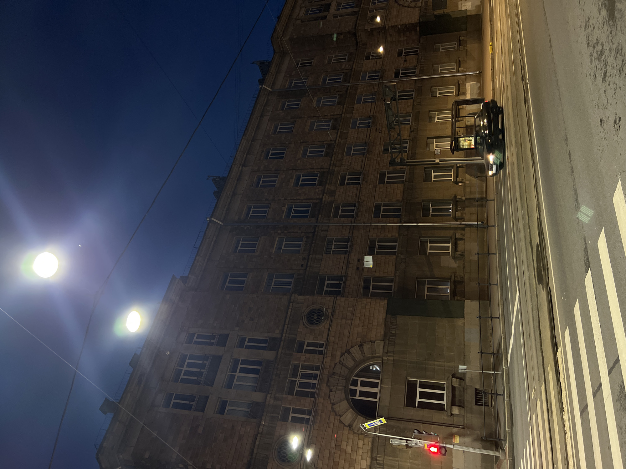 In the city of Unknown, Russia, at night, a wide, multi-lane street with tram tracks runs in front of a large, stone building. The building is illuminated by overhead streetlights, casting a warm glow on its many windows. The sky is a deep blue, suggesting late evening or early morning. A black car is driving on the street, its headlights on. Pedestrian crossings are visible on the road. A traffic light is red. A bus stop shelter is present on the sidewalk. The scene is devoid of people, creating a quiet, urban atmosphere.