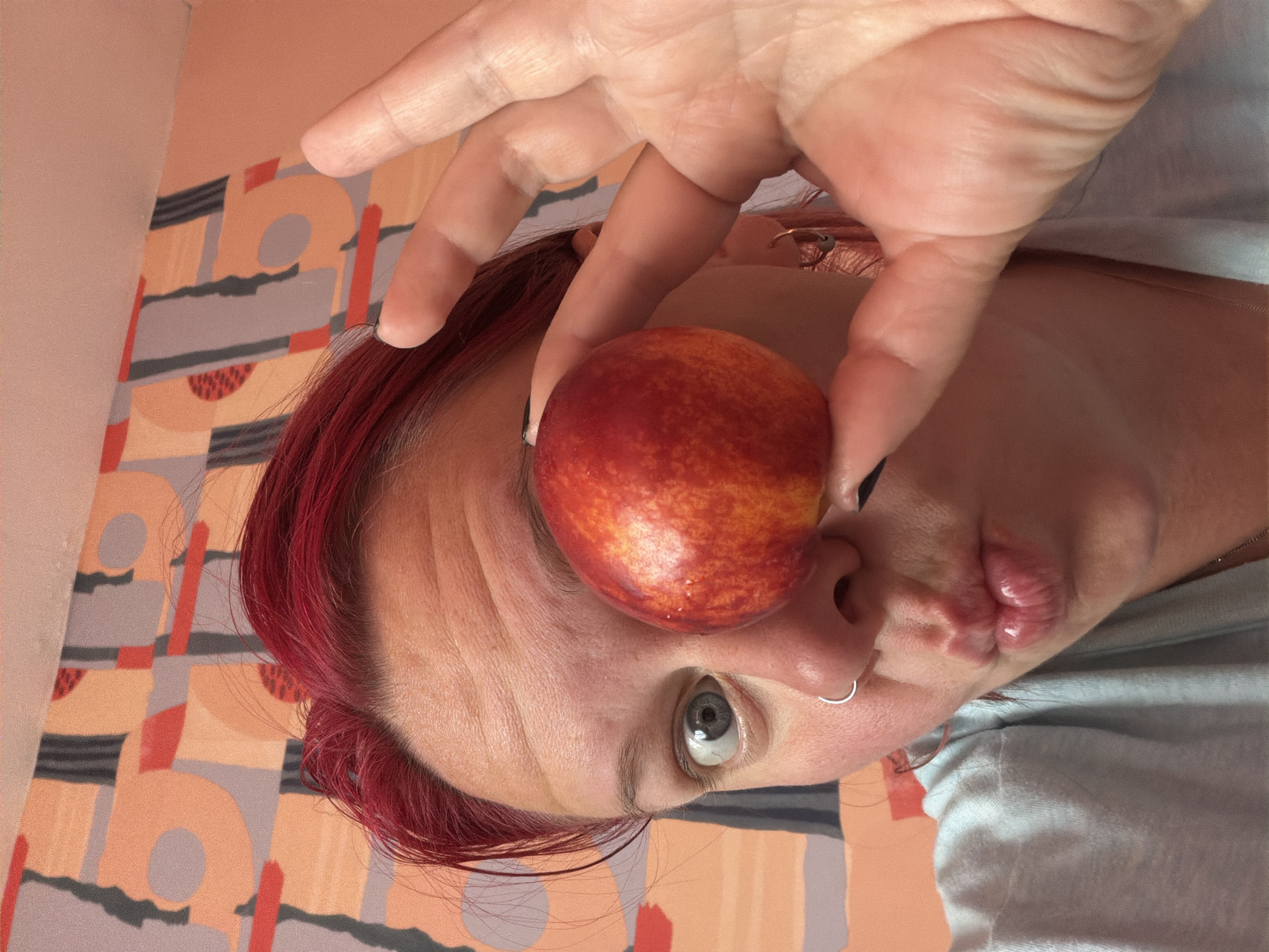 A person with bright red hair is holding a nectarine in front of their right eye, obscuring it from view. Their left eye is wide open and looking directly at the camera. Their lips are pursed as if to kiss. The person is wearing a light blue t-shirt and a silver nose ring. The background appears to be a wall with a colorful abstract pattern. The scene is brightly lit, possibly with natural light. There is no indication of the location being in Chaikovsky, Russia.