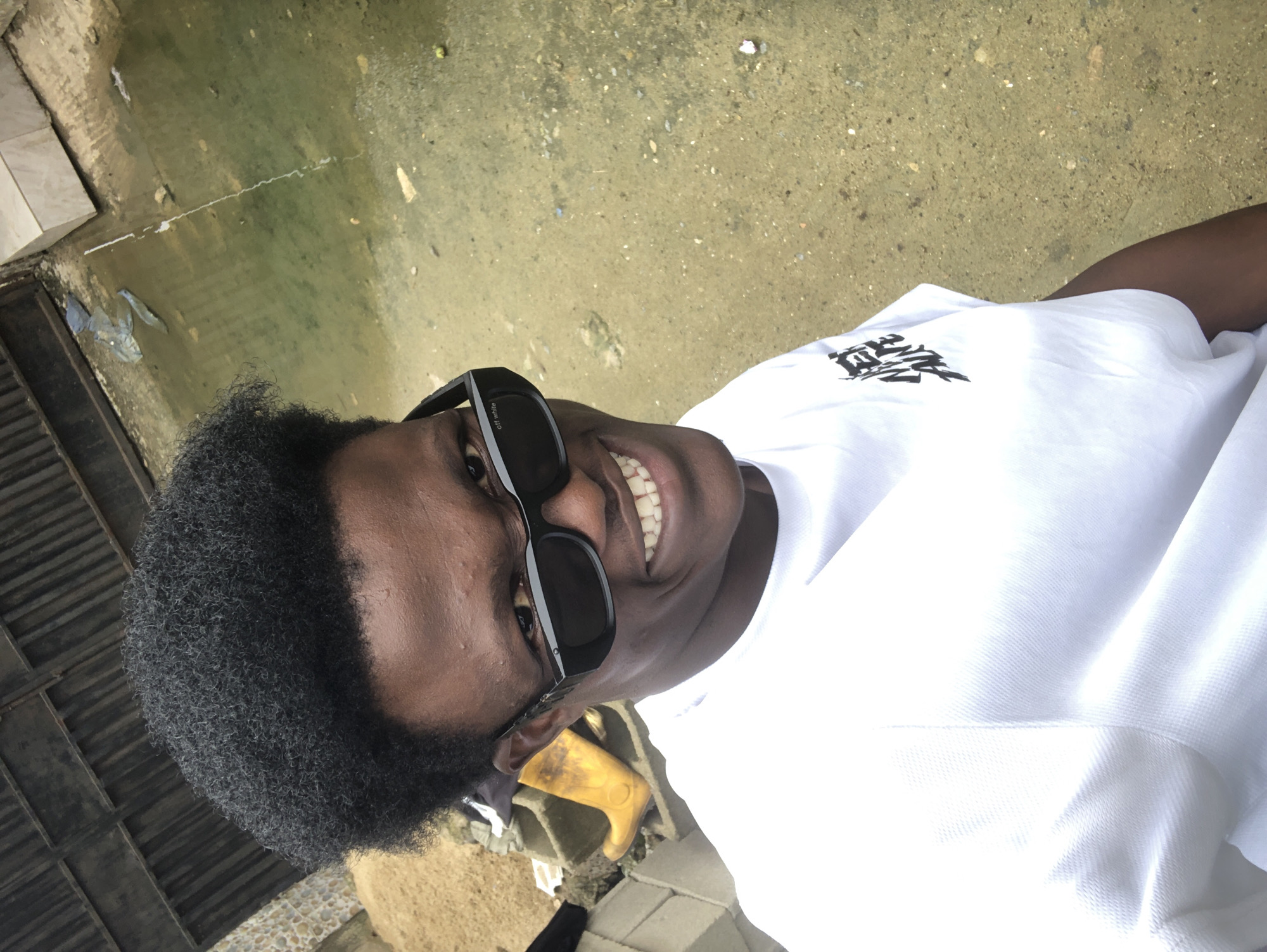 A medium shot captures a Black male posing for a selfie. He is wearing sunglasses and a white t-shirt with a graphic design on the left side of his chest. He has a playful expression, with his mouth open and teeth visible. The background appears to be an outdoor urban setting in Ado, Nigeria, possibly a back alley or a less-developed area. To the left, there are stacked concrete blocks and a bright yellow boot. Behind him, a dark metal fence or gate is visible, and beyond that, a body of murky, greenish water with some debris floating on the surface. The ground is sandy and uneven, with scattered small stones and dirt. The lighting suggests it is daytime, likely overcast or late afternoon, as the light is diffused.