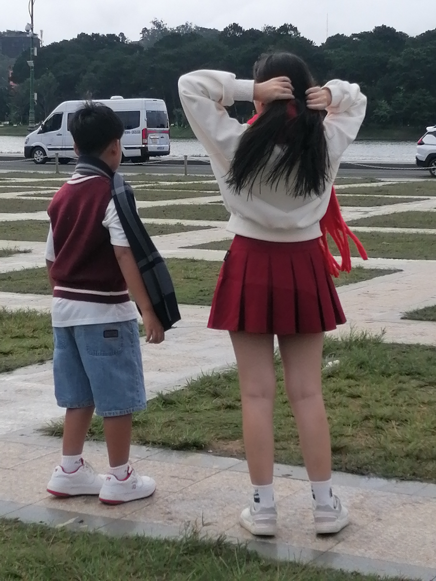 In Da Lat, Vietnam, two individuals, a young boy and a teenage girl, stand on a paved walkway adjacent to a grassy area. The boy, facing away from the camera, wears a maroon vest over a white t-shirt and denim shorts. A checkered scarf is draped over his left shoulder. The girl, also facing away, has dark hair and is tying it up. She is wearing a white sweater, a red pleated skirt, and white socks with sneakers. In the background, a white van is parked on a road that runs parallel to a body of water, beyond which lies a dense forest. The sky is overcast. The walkway appears to be part of a park or public area, indicated by the patterned paving stones and the surrounding greenery.