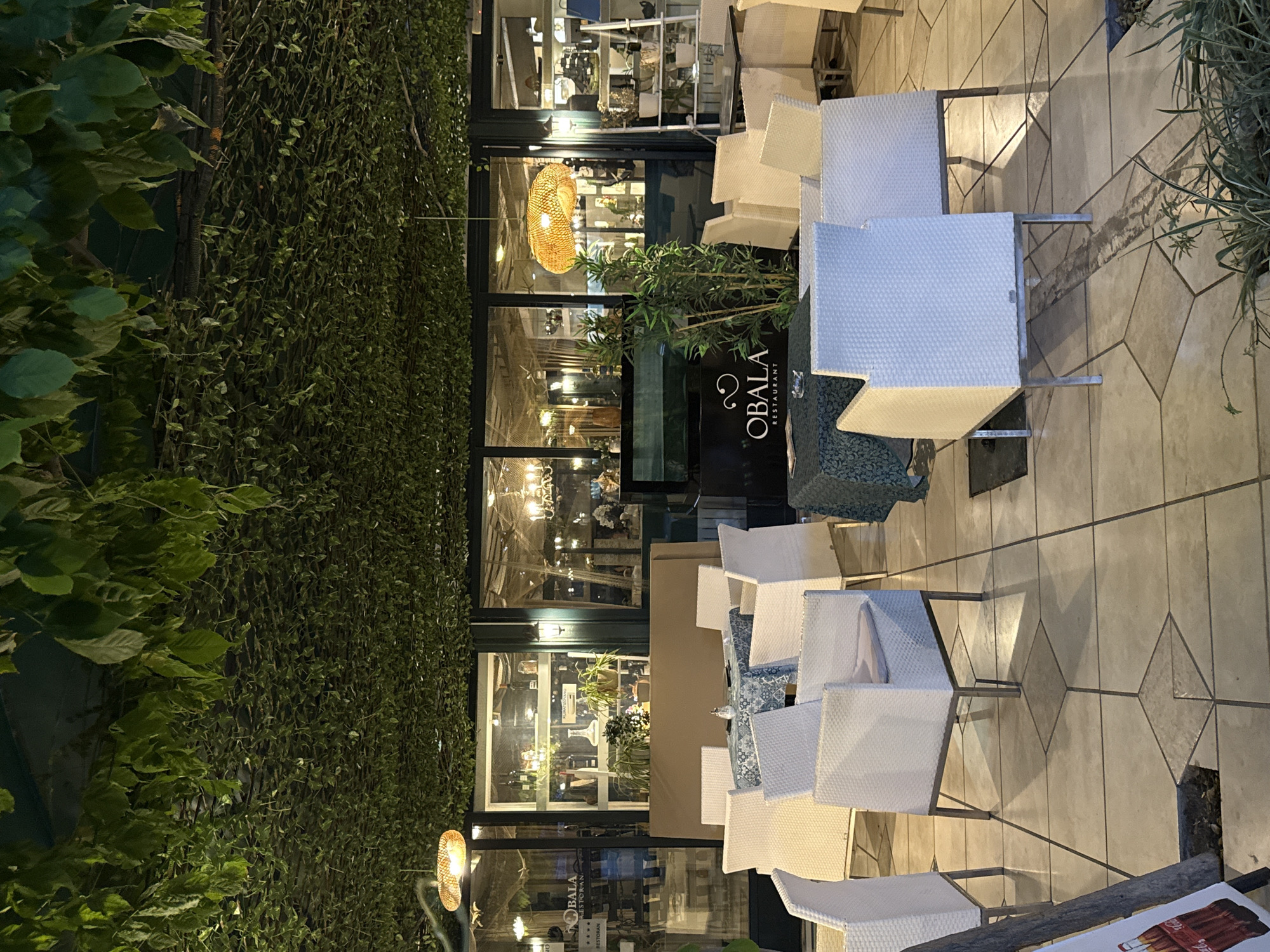 This media file displays an outdoor seating area of a restaurant at dusk. The scene is dominated by a large wall covered in lush green ivy on the left, with a paved patio area on the right filled with white wicker chairs and tables. The chairs are arranged around a table covered with a patterned tablecloth. Beyond the seating area, large glass windows reveal the interior of the restaurant, which is illuminated by warm lighting. Inside, shelves with decorative items and plants are visible. A prominent woven pendant light hangs in the window, casting a soft glow. The time of day appears to be evening, as indicated by the dim ambient light and the interior lighting of the restaurant. The weather seems calm, with no signs of rain or wind.
A black sign with white text is visible on the patio, reading "OBALA RESTAURANT". The paving stones on the patio have a geometric pattern of light and dark tiles. In the foreground, a partially visible piece of wood with some artwork on it rests on the ground. The overall impression is one of a tranquil and inviting dining setting, possibly in a coastal town given the location context of Budva, Montenegro.