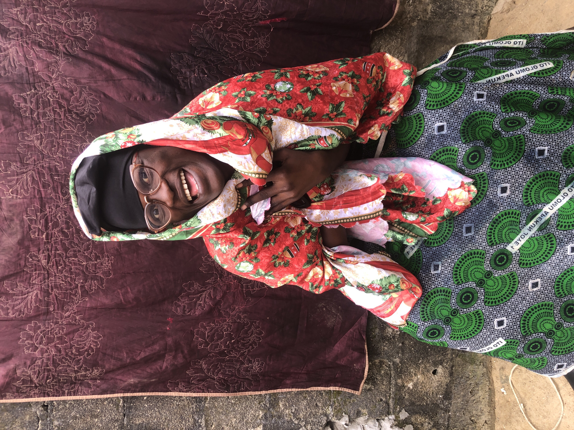 A Black woman with glasses and a headscarf is sitting in front of a dark red tapestry with a floral pattern. She is wearing a colorful, patterned outfit. The tapestry is hung on a concrete wall. The woman is smiling broadly and has her hands on her chest. The image appears to have been taken outdoors in Ado, Nigeria.