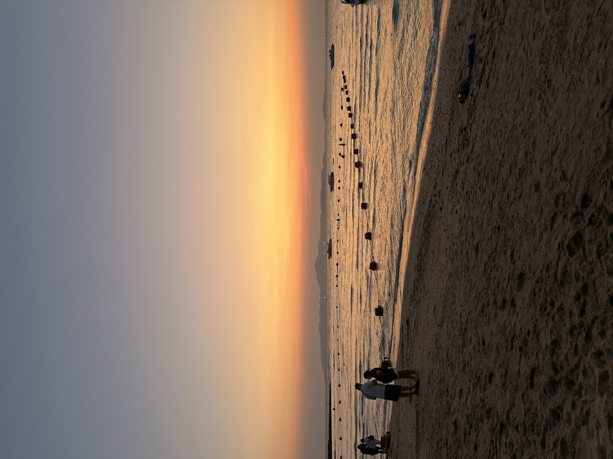 A landscape photograph, taken at dusk in Pattaya City, Thailand, depicts a beach scene. The sky transitions from a pale blue-gray on the left to a vibrant gradient of yellow, orange, and red on the right. The sea, reflecting the sunset's colors, is separated from the beach by a line of gentle waves. A series of dark, regularly spaced buoys or posts stretch into the water, forming a line parallel to the shore. Several boats are visible in the distance. On the right side of the image, a sandy beach is visible. Silhouetted figures of people stand near the water's edge in the foreground.