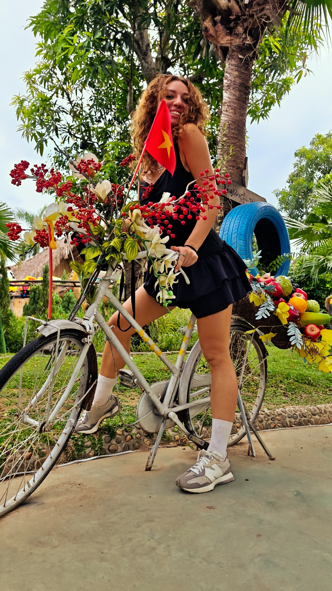 A woman with curly brown hair poses on a white bicycle in Hoa Lư Ward, Vietnam. She is wearing a black dress and white socks with sneakers. The bicycle is decorated with a large bouquet of red berries and white flowers in the front basket and a basket of colorful fruits at the back. A small Vietnamese flag is held up by the woman. The background features lush green trees and foliage, with a clear sky above. A blue tire is visible on the right side of the frame. The scene appears to be outdoors in a tropical or subtropical environment.