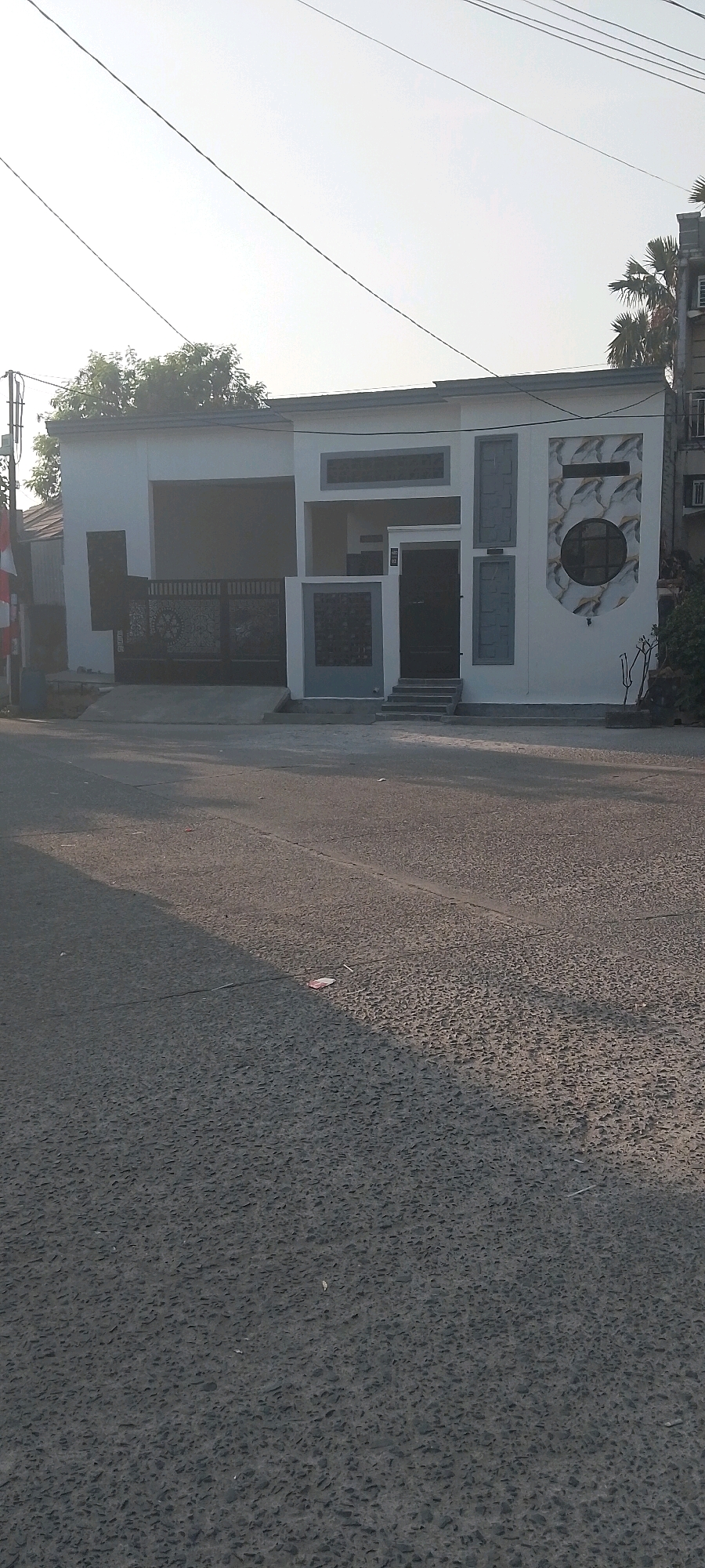 The image shows a modern, single-story building with a white facade, located on a street in Satriamekar, Indonesia. The building features a prominent dark metal gate, a dark-colored entrance door with steps leading up to it, and several decorative panels. One panel on the right side of the building has a circular design with marble-like patterns. Trees and palm trees are visible in the background, suggesting a tropical or subtropical climate. The street in front of the building is paved with asphalt and shows some signs of wear. Overhead power lines are visible against the bright sky. The lighting indicates it is likely daytime, with the sun casting shadows. There are no people visible in the image. The overall impression is of a residential or commercial property in an urban or suburban setting.