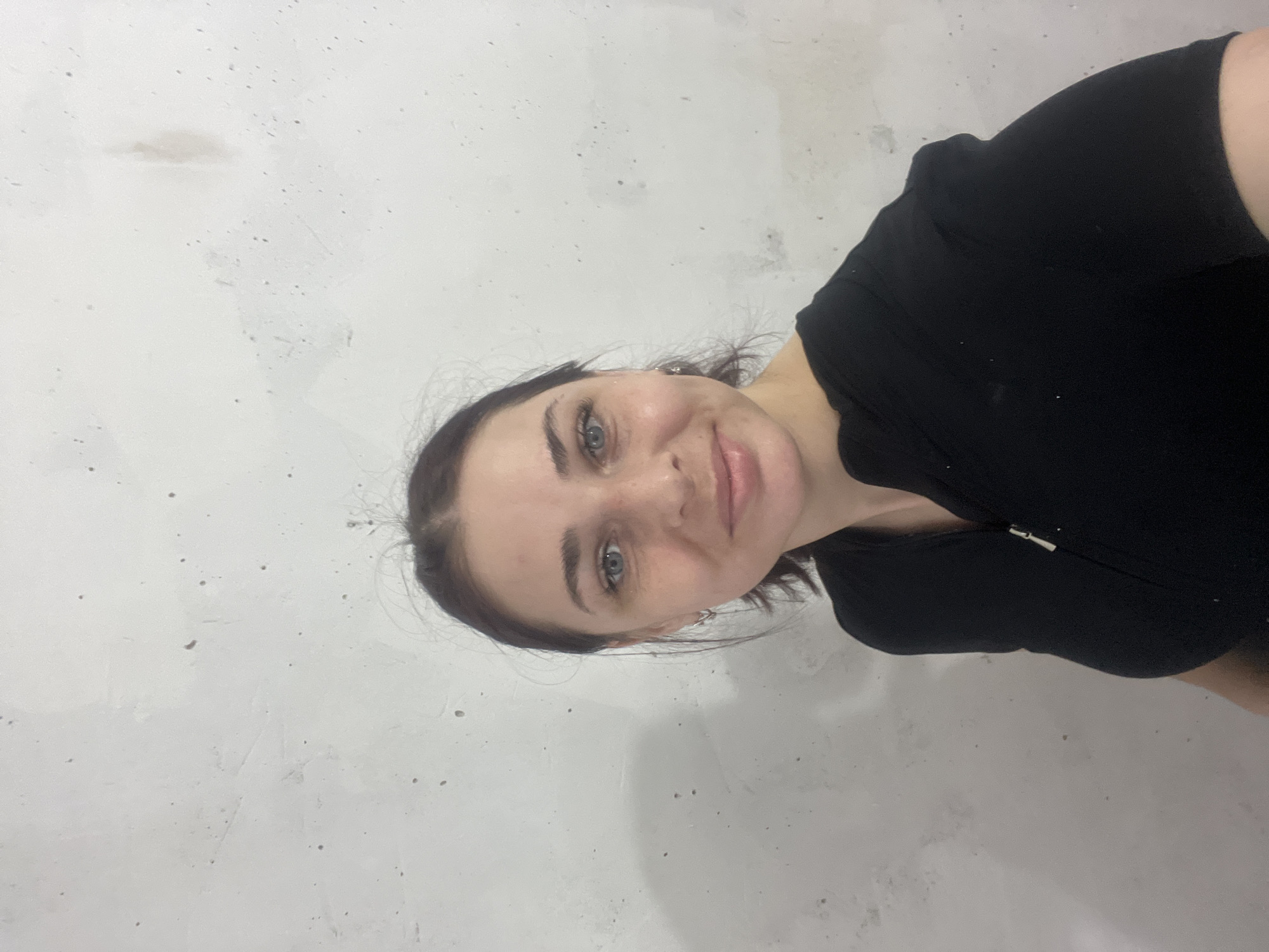 A woman with fair skin, blue eyes, and dark hair pulled back from her face is shown in a close-up shot. She is wearing a black top. The background consists of a light-gray wall with textured imperfections. There is no discernible information in the image to suggest a location in Chaikovsky, Russia, or any other specific place. The image provides no information about actions, interactions, or other subjects.