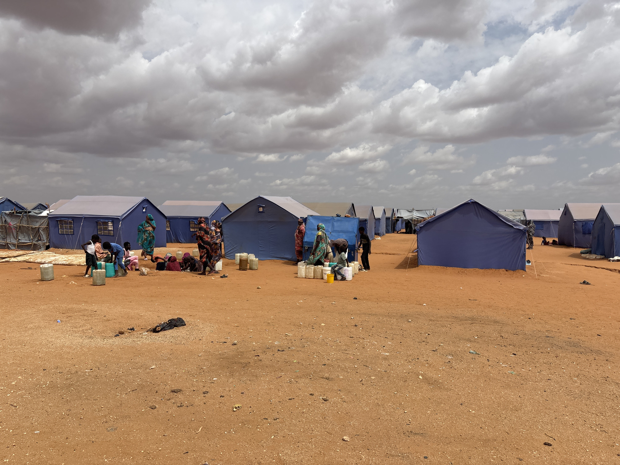 The image depicts a refugee camp in El-Obeid, Sudan, under a cloudy sky. A series of blue tents are set up on a reddish-brown, dusty ground. In the foreground and mid-ground, several people, primarily women and children, are engaged in various activities. Some are gathered around large plastic water containers, filling smaller buckets or jugs, suggesting water distribution or collection. Others are standing or sitting near the tents. The ground is littered with scattered debris, including plastic bottles and what appears to be a black bundle. The overall scene conveys a sense of temporary settlement and daily life in a humanitarian context.