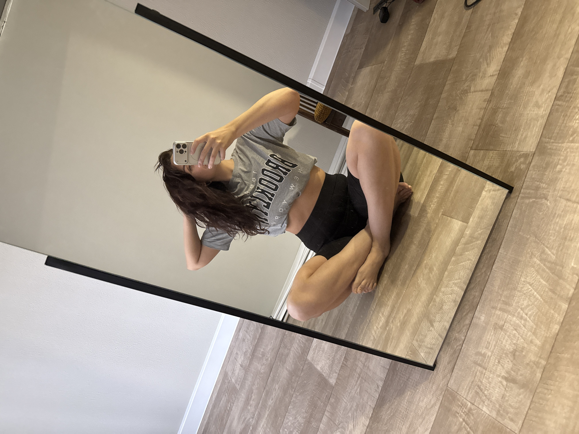 A mirrored image captures a woman with long, brown curly hair, clad in a gray crop top with "BROOKLYN" printed on it and black shorts. She is crouching on the floor, using her right hand to hold a cell phone up towards the mirror, presumably taking a selfie. The setting appears to be indoors, with a gray wall visible in the mirror's reflection. Outside the mirror's frame, a light-colored hardwood floor extends into the room. The location in the city of Chaikovsky, Russia cannot be confirmed from this image.