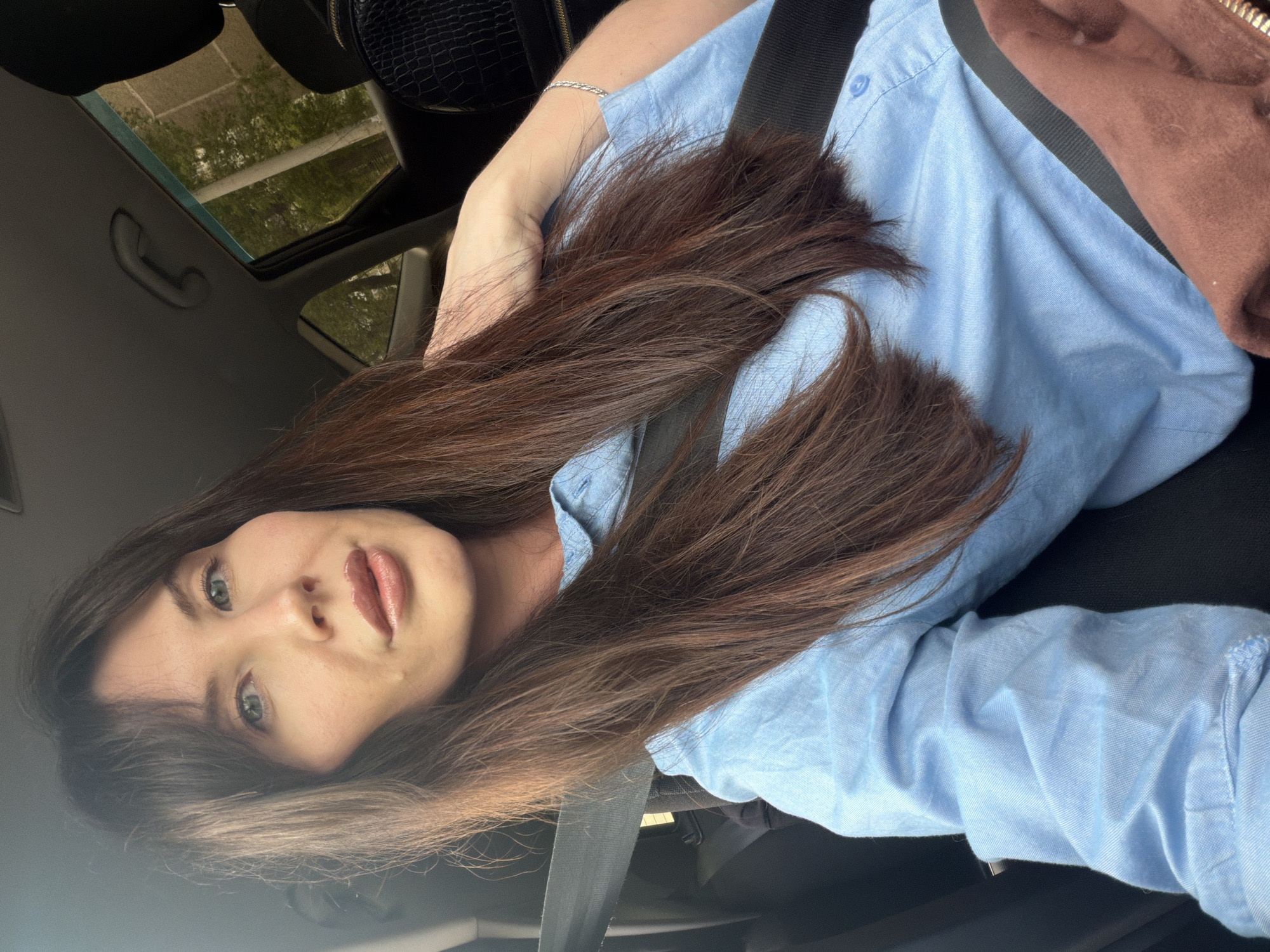 A woman with long brown hair and green eyes is seated in a car, wearing a light blue button-down shirt and a seatbelt. The location appears to be a car interior with a sunroof visible, suggesting a moving vehicle. Given the limited information, it is not possible to definitively confirm the location as Perm, Russia without additional evidence.
