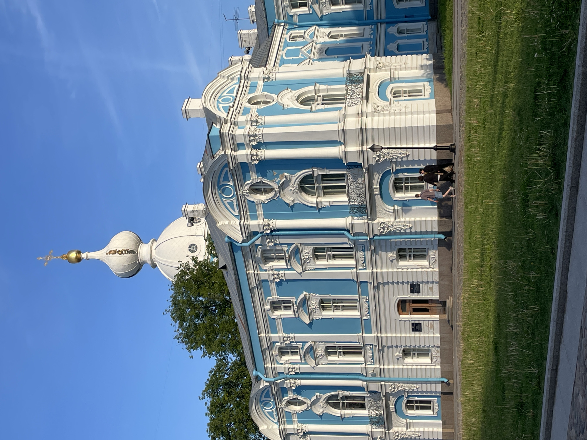 This image depicts a segment of the Smolny Cathedral and Monastery complex in Saint Petersburg, Russia, under a clear blue sky.
In the foreground, a multi-story building, part of the Smolny Monastery, presents a facade predominantly painted in a vibrant light blue, contrasted by white architectural details including columns, pilasters, window frames, cornices, and decorative mouldings. The lower portion of the facade is a light grey, above which the blue and white upper stories rise. Numerous windows, some arched and some rectangular, are visible, with several featuring decorative wrought-iron balconies. Blue downspouts run vertically along the facade. A wooden entrance door is centrally located on the ground floor, flanked by several dark rectangular signs.
In the background, positioned above and behind the foreground structure and partially obscured by green tree foliage, is the upper section of the Smolny Cathedral's white, onion-shaped dome. A golden orb, surmounted by a golden Orthodox cross, caps the dome.
On the paved walkway in front of the monastery building, three individuals are walking from left to right. The person on the far left wears a light-colored shirt and shorts. The middle individual is dressed in a dark top and trousers, while the person on the right wears all black attire. A black lamppost stands on the pavement to the right of the walking group.
The immediate foreground consists of a vibrant green grassy area, bordered by a grey concrete curb and a darker paved surface at the very bottom of the frame. The sky is bright and clear, displaying a uniform light blue hue, with a faint linear contrail visible in the upper right quadrant. Shadows are visible on the ground and parts of the building, indicating direct sunlight from the left.