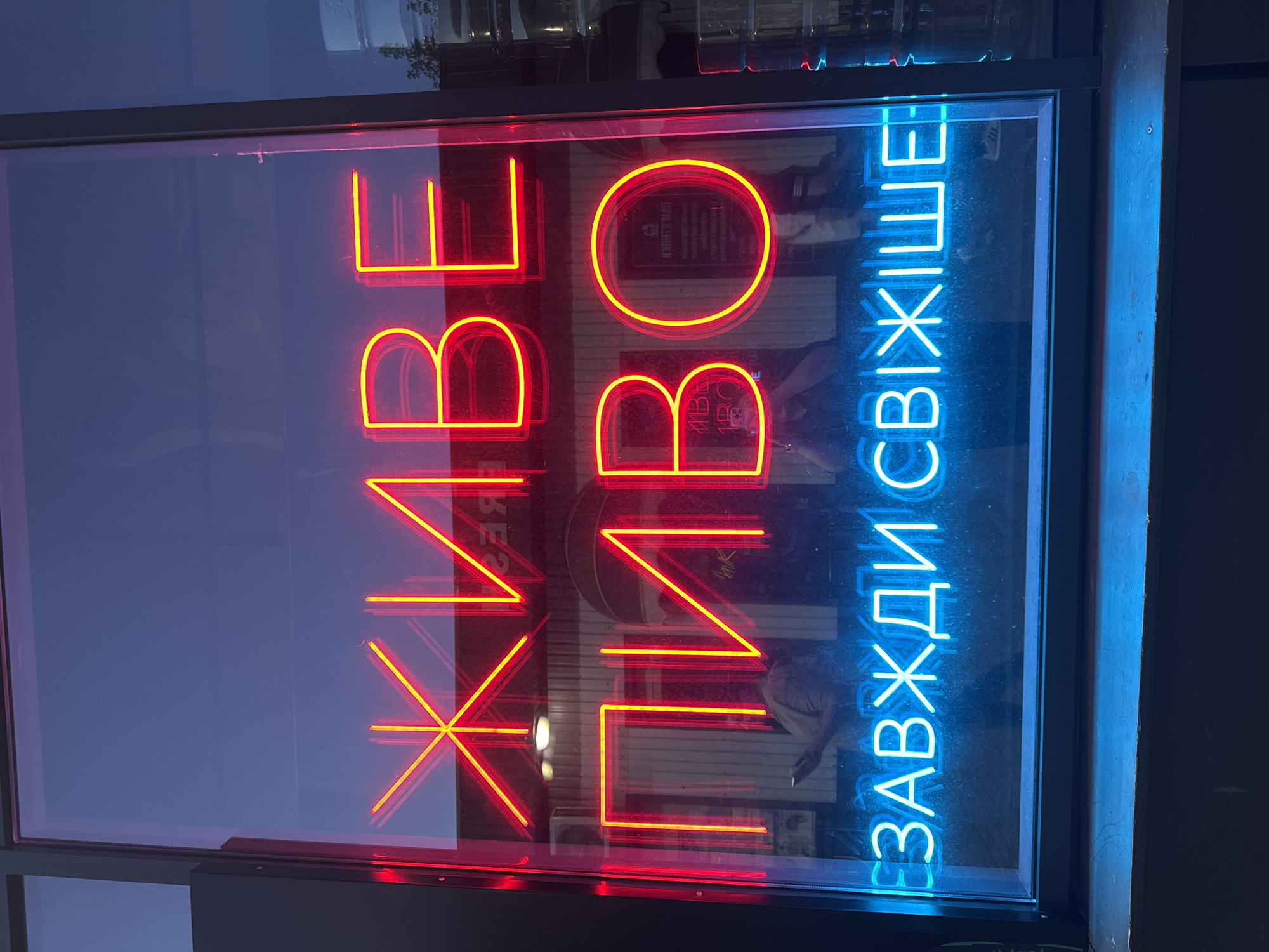 The image shows a neon sign in Ukrainian that reads "ЖИВЕ ПИВО ЗАВЖДИ СВІЖІШЕ" which translates to "LIVE BEER ALWAYS FRESHER". The sign is in red and yellow neon for the top two lines and blue neon for the bottom line. The sign is displayed in a window, likely of a bar or establishment, and is reflected in the glass along with the dim interior. The exterior environment suggests it is nighttime due to the dark sky and the illuminated sign. The location is identified as Kremenchuk, Ukraine. There are reflections of people in the glass, some holding phones, indicating they are likely indoors or passing by the window. The window itself is framed by dark material, and a dark ledge or sill is visible below the window.