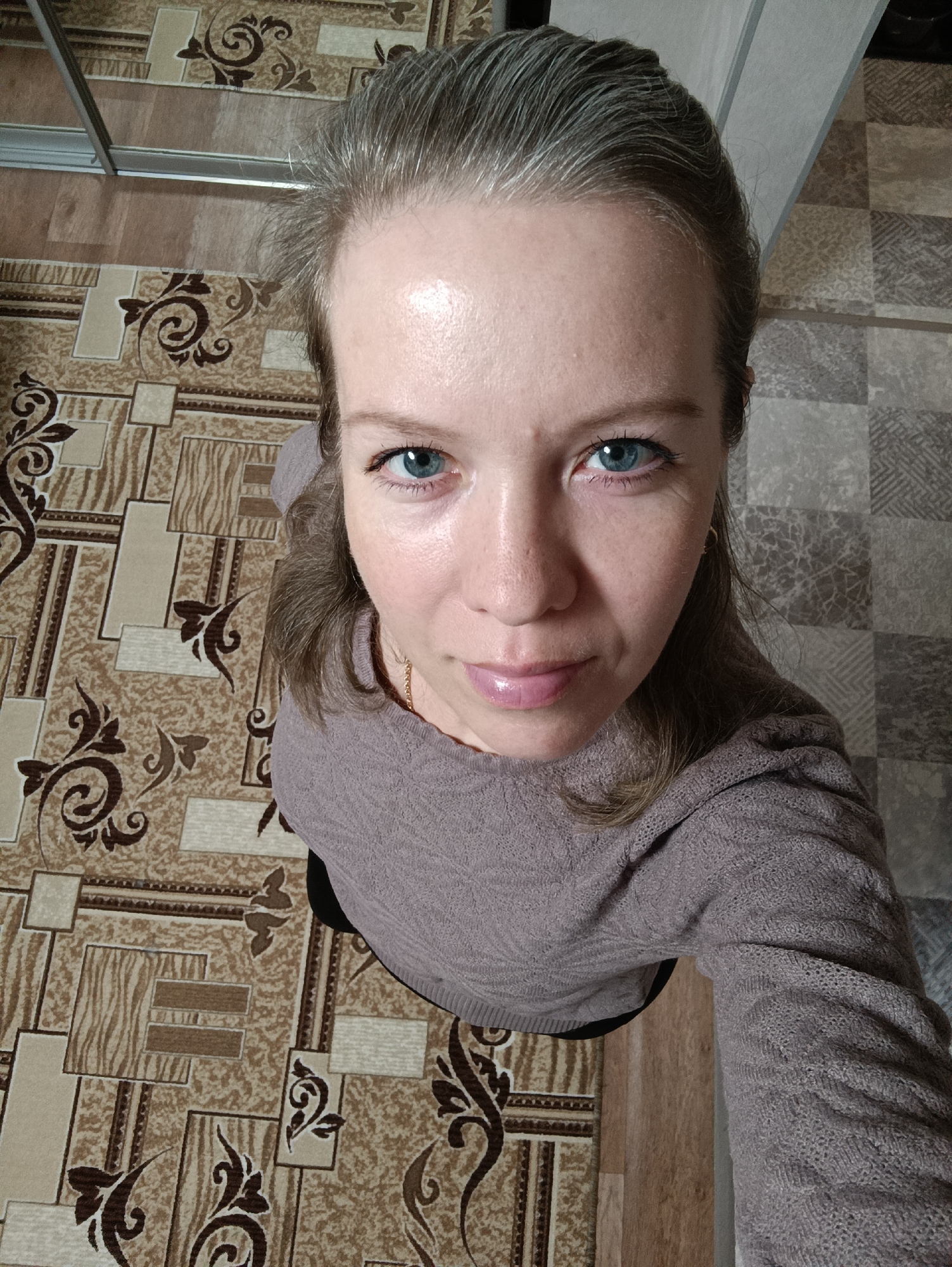 A woman with fair skin, blue eyes, and graying hair looks directly at the camera from a low angle. She wears a mauve-colored sweater and a gold chain necklace. The background shows two different types of flooring, one with a patterned carpet and the other with a square-tile pattern. The image was likely taken indoors. While the image was taken in Chaikovsky, Russia, there is nothing in the image to suggest a specific location in Chaikovsky, Russia.