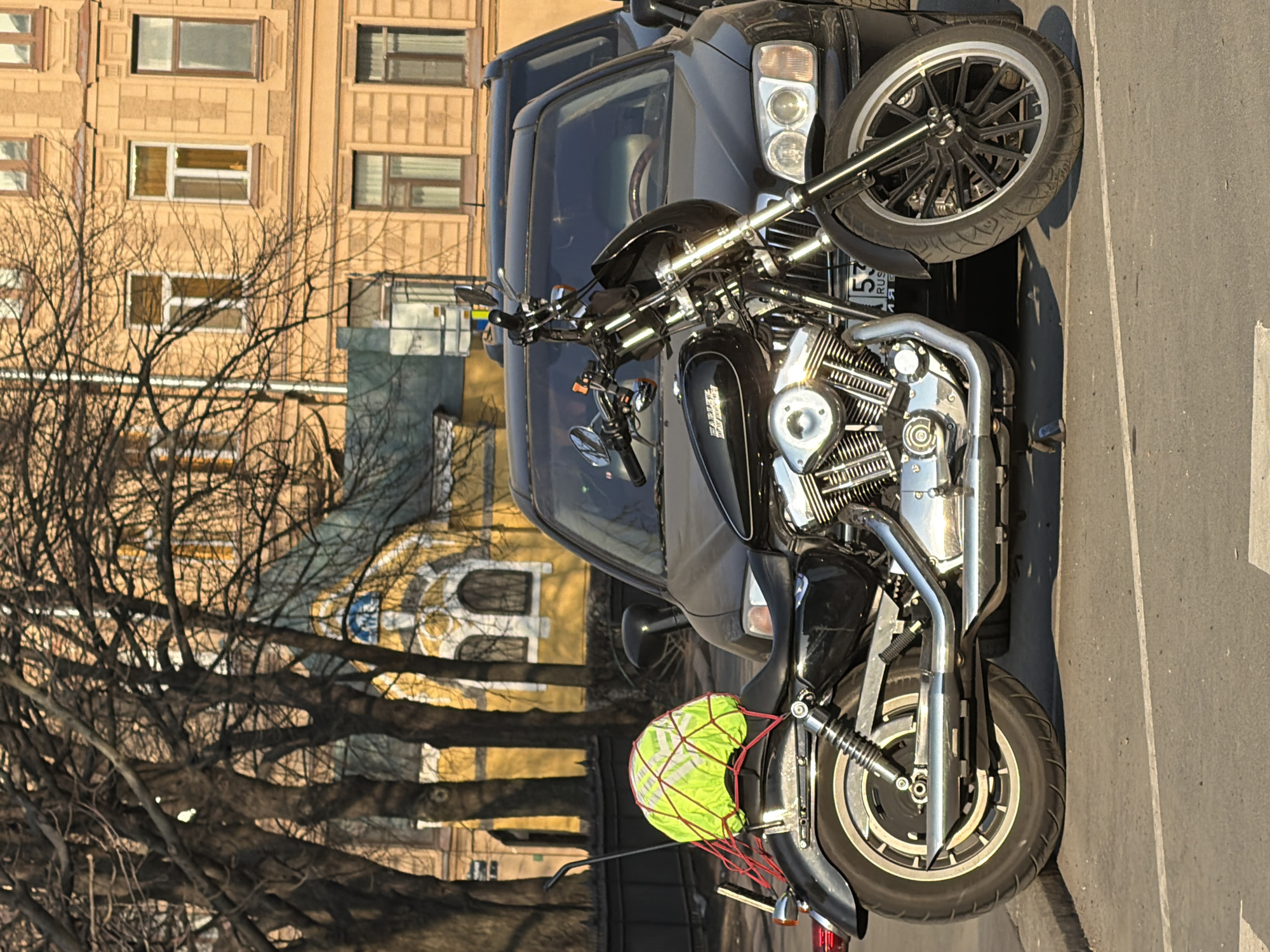 The image shows a black motorcycle parked next to a dark-colored SUV on a street in Saint Petersburg, Russia. The motorcycle has a bright green net attached to the back. Buildings are visible in the background, including a yellow building with a blue and white design and another beige building with window patterns. Bare trees are also visible. The road surface appears to be asphalt.