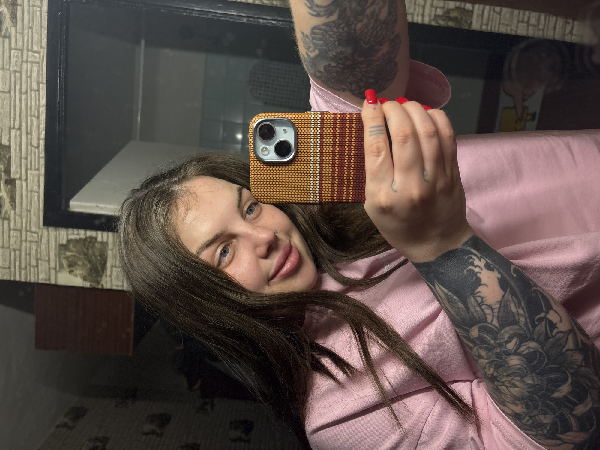 A woman with light skin, brown hair, and blue eyes is taking a selfie with a phone in a tan and maroon striped case. She is wearing a pink shirt and has tattoos on both arms, one showing a dragon and the other floral designs. The background shows a black-framed window with light reflected in it, and a patterned wall. The woman appears to have a nose piercing. Based on the prompt, the location is Chornomorskoe, Ukraine.
