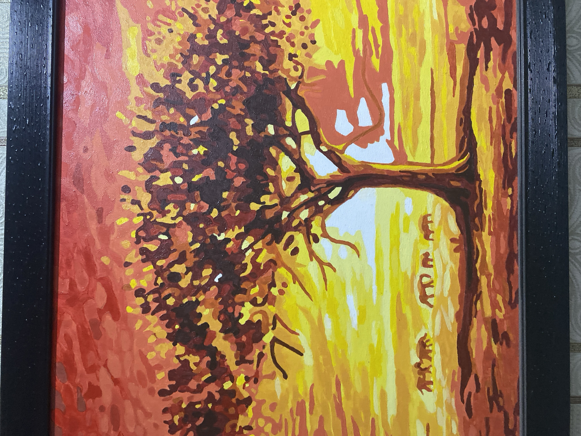 The image is a painting of an African savanna landscape during sunset. A large, silhouette tree dominates the center of the frame, its branches spread wide against a vibrant sky of red, orange, and yellow hues. In the distance, a herd of elephants is depicted walking across the plains, casting long shadows. The painting style is stylized and graphic, with bold brushstrokes and a limited color palette. The painting is framed by a black, textured border, suggesting it is a finished artwork ready for display. There is no indication or information within the image itself that connects it to Chernogorsk, Russia, or any other specific city or location.