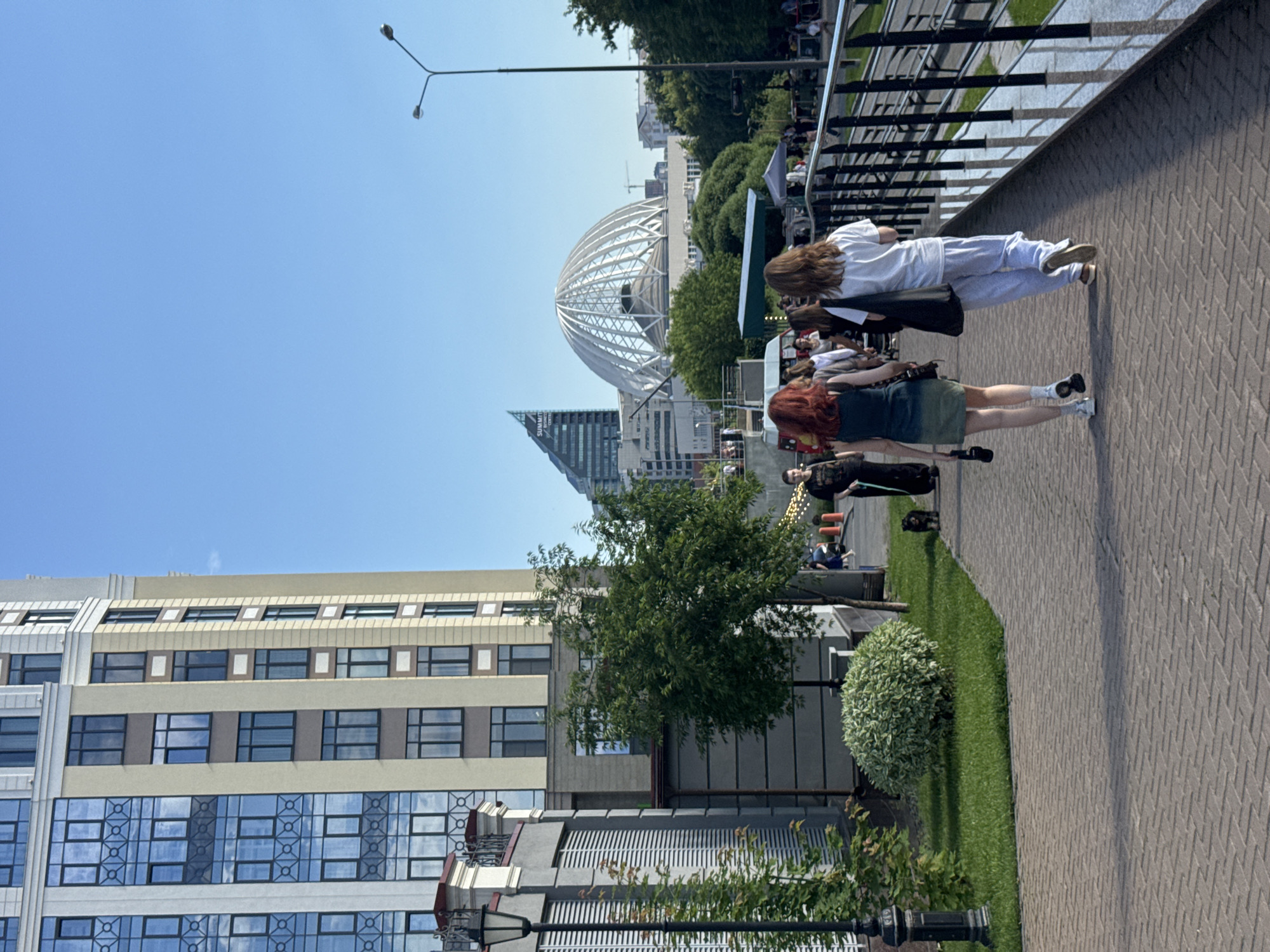This media file captures an outdoor scene in Yekaterinburg, Russia, under a clear blue sky, suggesting a bright, sunny day. The image is oriented vertically, with the sky occupying the upper portion and the ground below.
The primary subjects are several people walking along a paved pathway that runs alongside a grassy area. Two young women are prominently featured in the foreground, walking away from the viewer. One wears a white top and light-colored pants, while the other is in a dark green skirt and white top. Behind them, other individuals and a small black dog are visible, suggesting a public space with some foot traffic.
In the background, a striking modern architectural structure with a domed, lattice-like roof is visible, characteristic of some exhibition centers or event venues. Adjacent to this are modern high-rise buildings, indicating an urban environment. The pathway is made of light-colored brick pavers laid in a herringbone pattern. Adjacent to the path is a neatly mowed lawn with a meticulously shaped spherical shrub.
A streetlamp is visible in the upper left corner, and a railing runs along the edge of a raised area on the left side of the path, possibly a balcony or elevated walkway. The overall impression is one of a pleasant day in a modern city with public spaces designed for leisure and pedestrian activity. No discernible text is visible in the image.