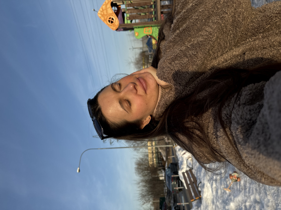 A female individual is depicted in a close-up, slightly low-angle shot, facing forward with her eyes closed and a serene expression, her chin resting on her right hand. She has long, dark hair, with black sunglasses perched on her head, and is wearing a brown textured outerwear piece over a light brown turtleneck. The right side of her face is illuminated by direct sunlight, casting a warm glow.
The scene is set outdoors in Akulovo (Gp Kubinka), Russia, on a clear, sunny day during winter. Snow covers the ground in the foreground and midground. To the left, a wooden park bench with light-colored slats is partially covered in snow. Beyond the bench, a paved area or street contains several parked vehicles, including a dark-colored sedan. White bollards or posts delineate the edge of the paved surface. Bare deciduous trees are visible in the background on the left.
To the right, a multi-colored children's playground structure, primarily orange, green, and purple, stands on the snow-covered ground. A street lamppost with a white fixture and a power line pylon are visible against the clear blue sky in the upper background. Overhead power lines traverse the sky. Distant structures and additional bare trees are present on the horizon. A small, orange and white toy is partially visible in the snow in the lower-left foreground.