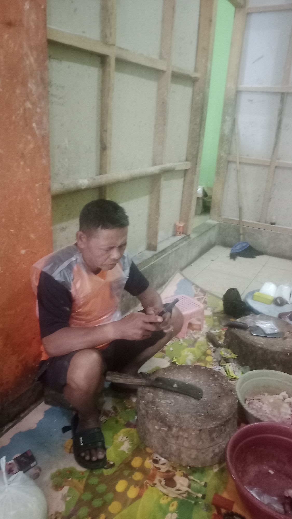 A man sits on the floor in a dimly lit room, wearing a black and orange t-shirt and dark shorts. He is wearing black sandals. The man is holding a mobile phone in his right hand and appears to be looking at it intently. In front of him, on a colorful mat, is a large, round stone mortar with a knife resting on it. To the right of the mortar is a red bowl containing what appears to be minced meat. Behind the man, a wooden partition divides the room, with a green wall visible in the background. The overall impression is that of a preparation area, possibly for food. The location is a domestic setting in Karyamandala, Indonesia, suggesting a workshop or kitchen area.