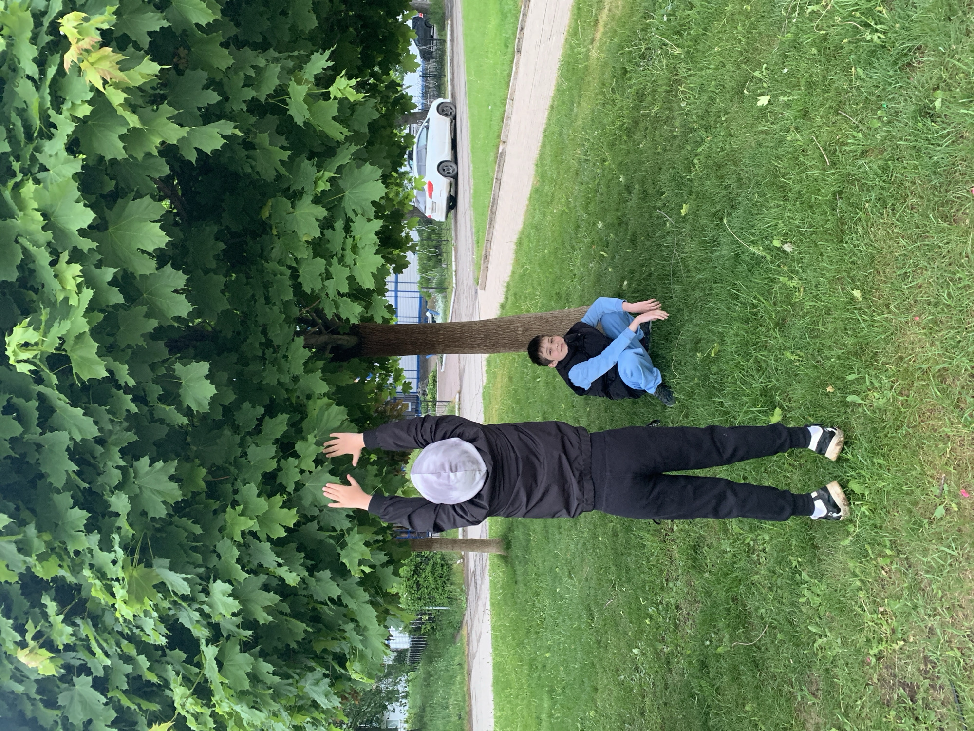 A horizontal outdoor shot depicts two people, one lying on the grass with a gray and white beanie, black jacket and pants, and black and white shoes, arms extended touching tree leaves. The other person is sitting with legs crossed, wearing blue pants, a blue shirt, and a black vest. A tree trunk is visible in the middle ground. A sidewalk runs parallel to the grass, with a parked white car visible in the background. The scene is set in a grassy area in what appears to be a park or residential area. Based on context, the location is Chaikovsky, Russia.