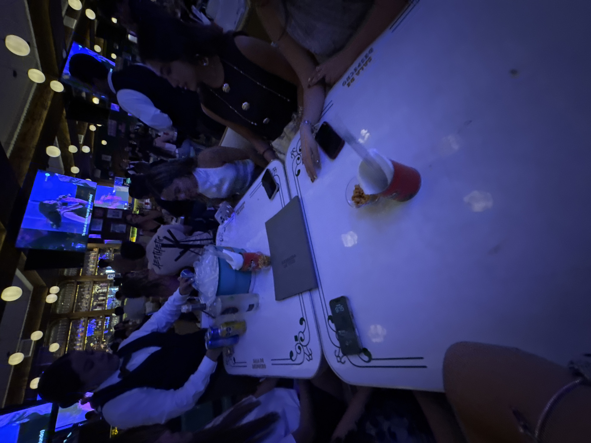 The image displays an indoor night scene within a bar or restaurant, identified as being in the city of Puebla, Mexico. The environment is characterized by dim blue ambient lighting and circular yellow-orange ceiling fixtures. A long, multi-tiered bar stocked with bottles is visible in the background. Multiple large flat-screen televisions are mounted throughout the venue, displaying a music video featuring a female performer; Spanish text, "Te daré el último beso (el más profundo)", is partially legible on one screen.
In the mid-ground, a female server, wearing a white collared shirt, dark vest, and bowtie, is positioned left of center, attending to a white rectangular table with a dark, ornate border. Her left hand is near a blue bucket containing ice and wrapped items. On the table, alongside the ice bucket, are several beverage cans (one blue, one yellow), a clear liquor bottle, and a dark grey menu or binder.
Seated at the table are at least four female patrons. One woman in a white sleeveless top is visible centrally, facing right. To her right, another woman is seated, wearing a dark sleeveless top with prominent gold button-like embellishments and a long necklace. Her right hand, adorned with rings and a bracelet, rests on the table. The arm of a third woman, similarly adorned with jewelry, is visible on the far right. In the immediate foreground on the table, a red cup contains snacks, and a black smartphone lies face-up, displaying the time "9:15". Another red cup and a second smartphone, placed face-down, are also visible.
Numerous other patrons are present in the blurred background, indicating a populated social gathering. One male patron in the background wears a white t-shirt with "AESTHETIC" printed on the reverse.