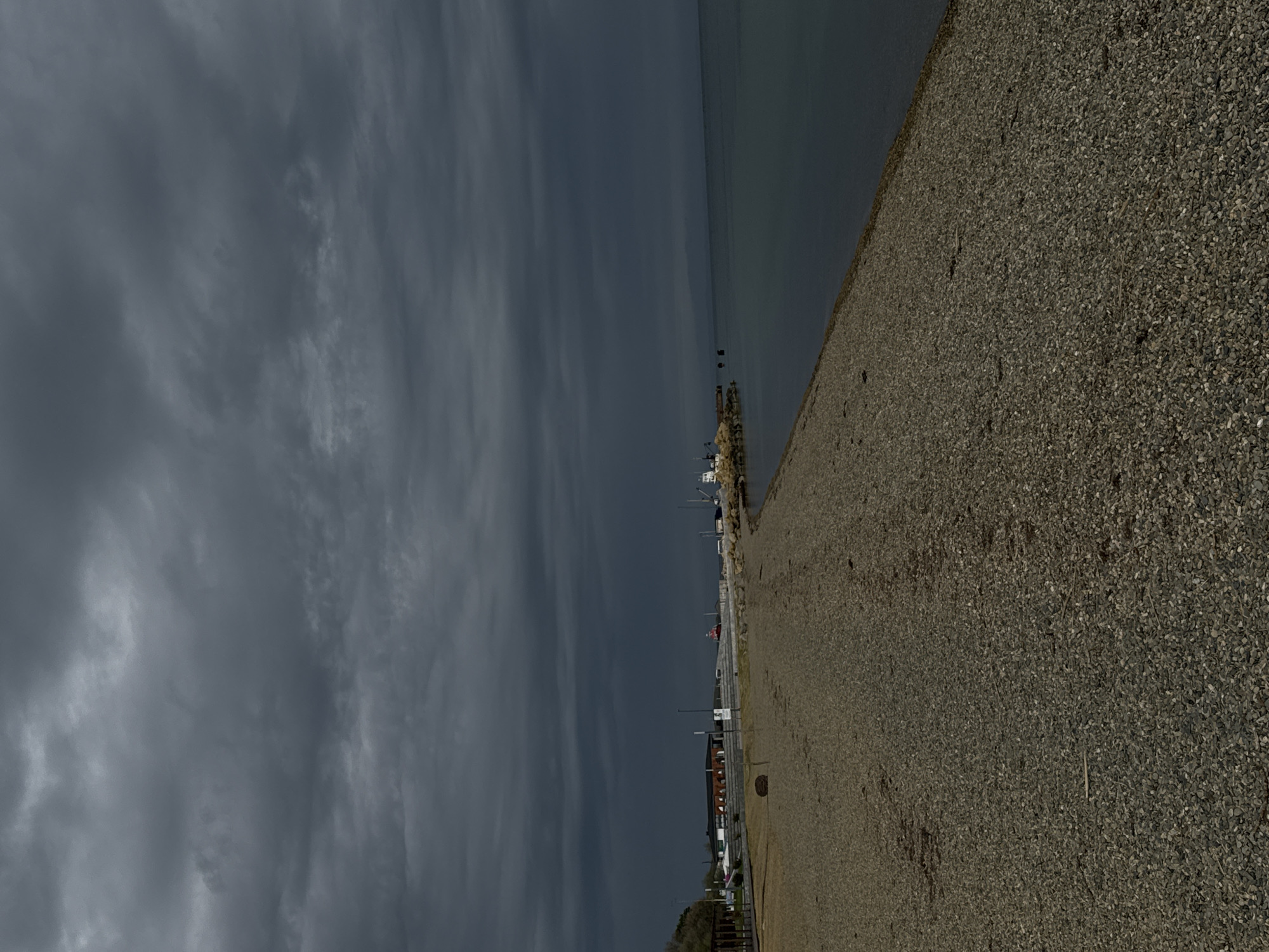 The image depicts a beach in Taman, Russia, under a somber, overcast sky. The scene is dominated by a shingle beach, with small, gray pebbles extending across the lower right half of the frame. Adjacent to the beach is a calm body of water, likely the Black Sea, its surface reflecting the dull sky. A pier or breakwater with various boats or structures is visible along the water's edge. Beyond the beach, buildings, lamp posts, and other urban elements suggest a small town or village. The sky is filled with dense, gray clouds, indicative of an approaching storm or persistent poor weather. There are no discernible people or specific actions captured in the image.