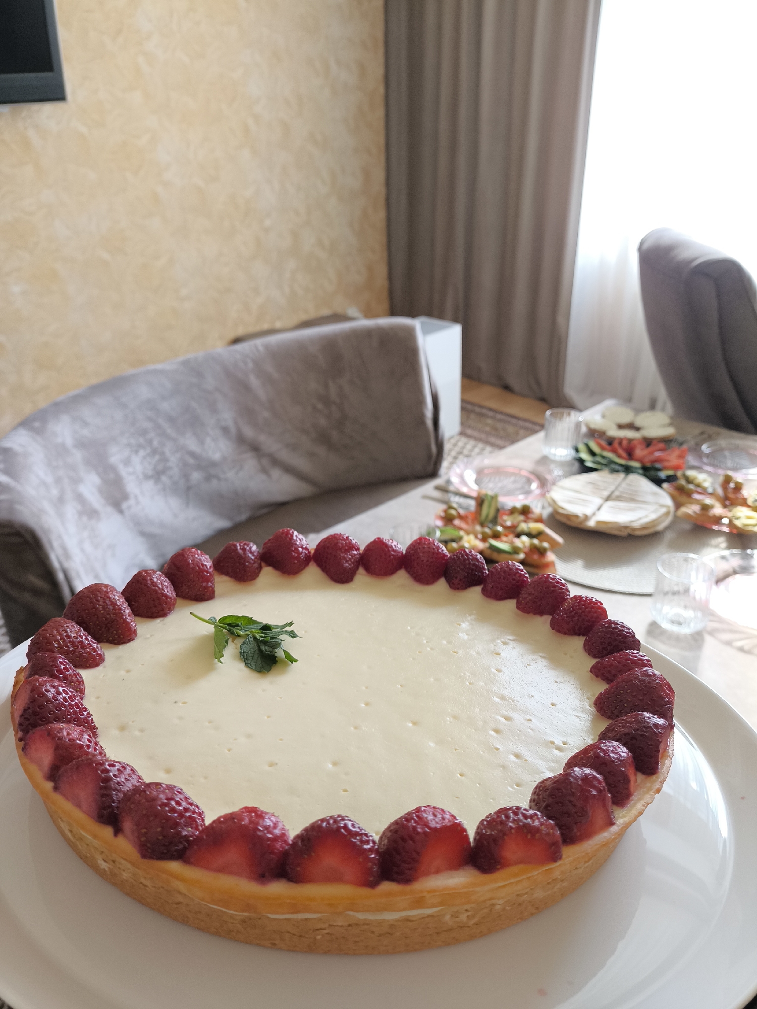 A close-up shot shows a strawberry cheesecake on a white plate, placed on a table. The cheesecake is round with a creamy yellow filling and is decorated with fresh strawberries around the edge, and a sprig of mint in the center. In the background, a couch covered with a grey blanket and a partially visible chair are present. The room has a textured yellow wall and light-colored curtains. Several small glasses and plates with food are visible on the table. The lighting suggests an indoor setting during the day.