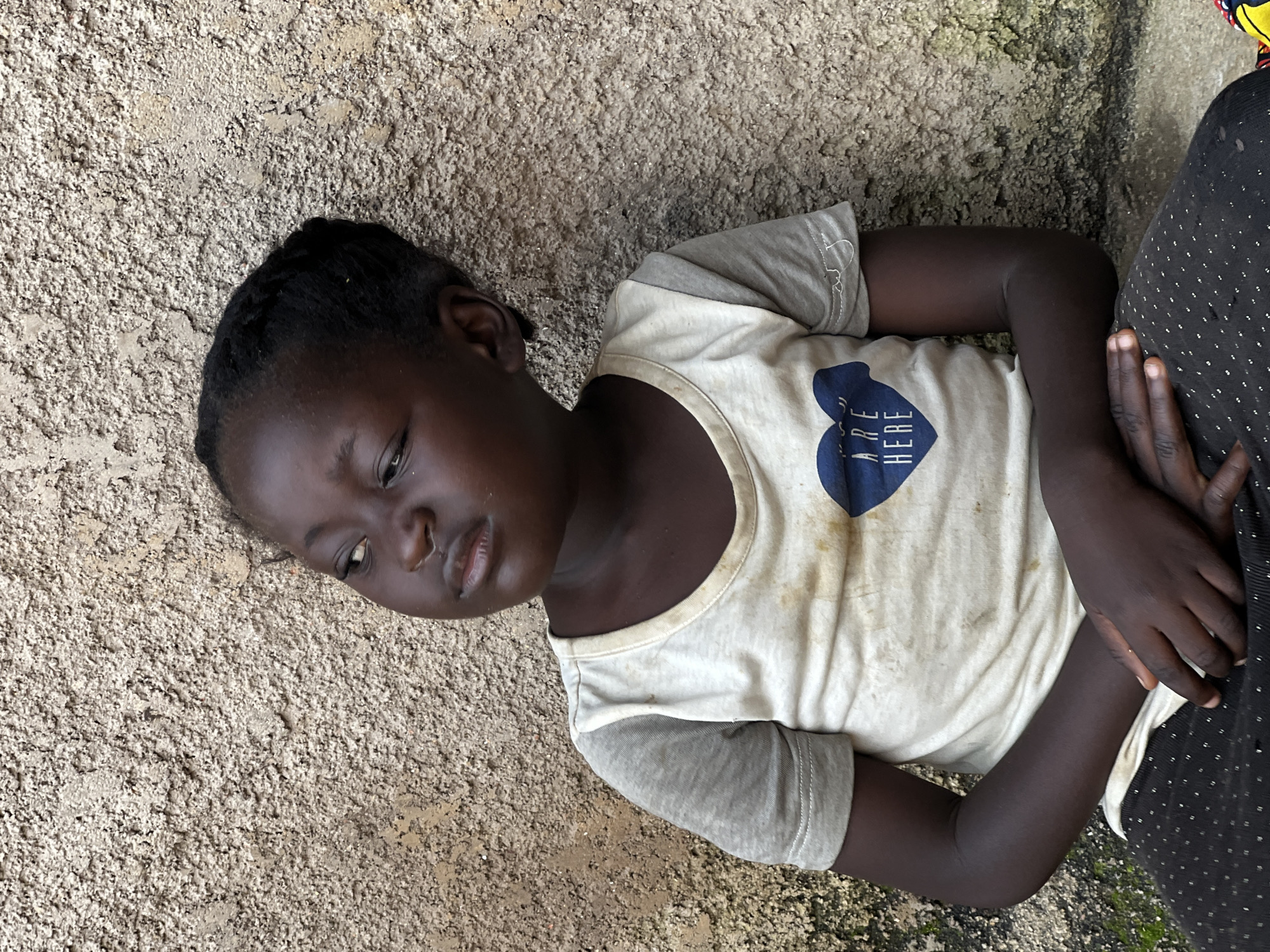 A young Black girl, with dark skin and braided hair, is shown from the chest up, seated against a rough, textured, light brown wall. She is looking to her left, away from the camera. She wears a light-colored t-shirt with grey sleeves. A dark blue heart graphic with the words "ARE HERE" is printed on the chest of the t-shirt. The t-shirt appears dirty and stained. Her hands are clasped in her lap. The background wall has patches of green moss. The scene appears to be outdoors, possibly in a residential area, given the texture of the wall and the presence of moss. The image was taken in the city of Jos, Nigeria.