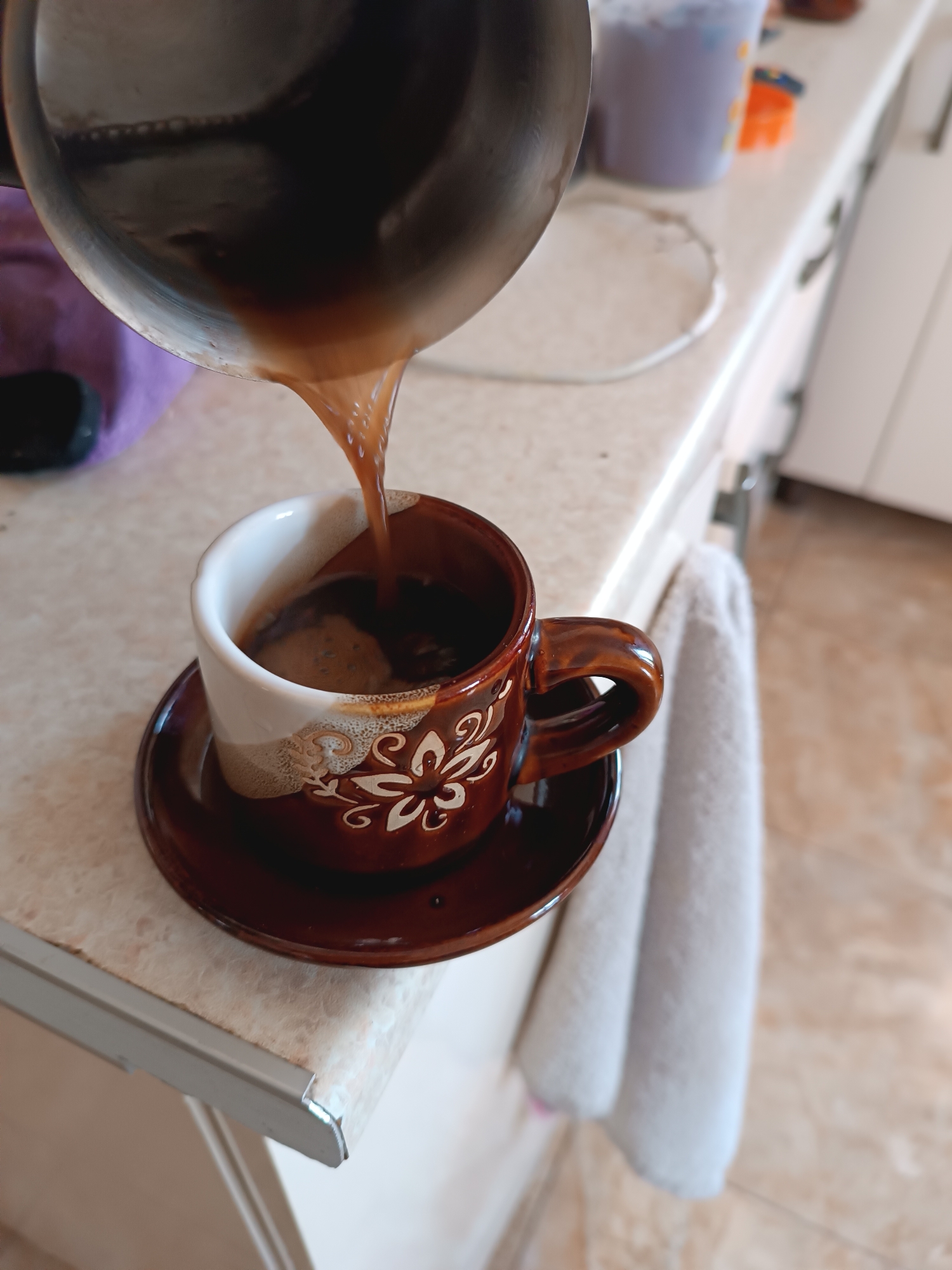 A high-angle medium shot shows coffee being poured from a metallic pot into a white and brown ceramic cup with a floral pattern. The coffee stream is visible as it enters the cup, which is sitting on a matching saucer. The scene takes place on a speckled countertop. A white towel hangs on the right side of the frame, and kitchen cabinets are visible in the background. The lighting is bright, suggesting an indoor setting during the daytime.