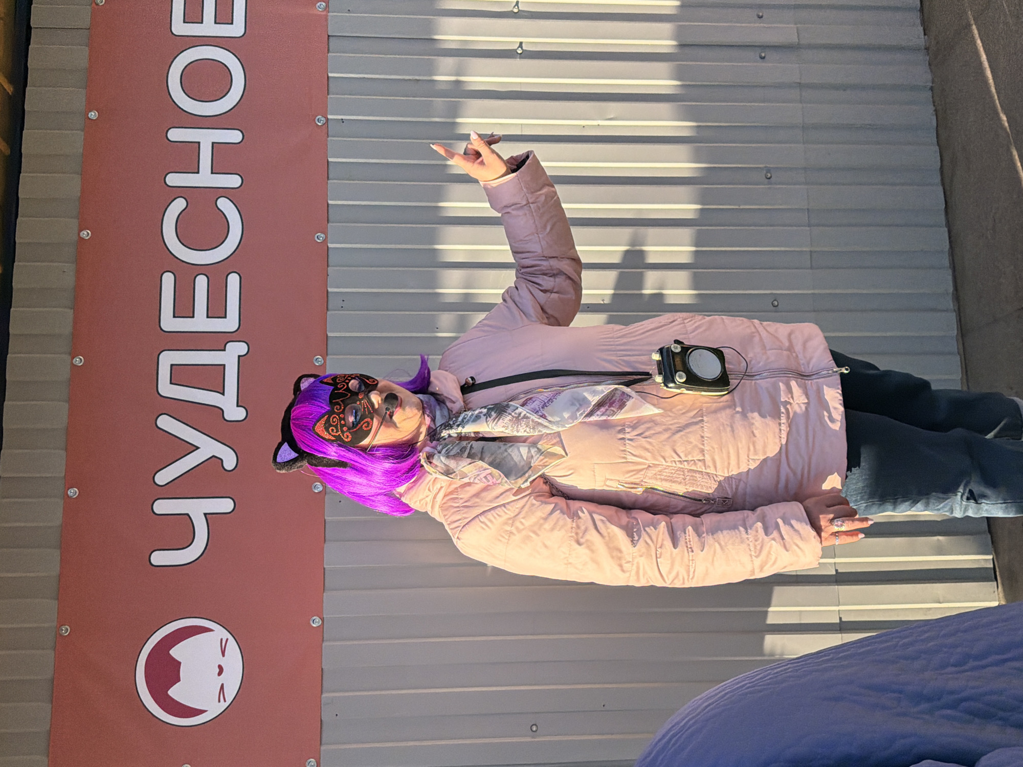 A person dressed in a costume stands outdoors in Saint Petersburg, Russia, against a corrugated metal wall. The individual is wearing a pink puffer jacket, dark blue pants, and a patterned scarf. Their head is adorned with a purple wig and black cat ears, while a black and gold cat mask covers their eyes and nose. The person's right arm is raised, with the hand making a gesture where the index and pinky fingers are extended. A small black electronic device hangs from their neck.
To the left of the person is a tall, vertical banner attached to the wall. The banner is reddish-brown with white text and a logo. The visible Russian word on the banner is "ЧУДЕСНОЕ" (CHUDESNOE), which translates to "WONDERFUL" or "MARVELOUS." Below the text, there's a circular logo featuring a stylized white cat's head with red outlines on a red background.
The scene is illuminated by bright daylight, casting distinct shadows on the corrugated wall, suggesting a sunny day. The pose and costume indicate a playful or expressive event, possibly related to cosplay or a themed gathering.