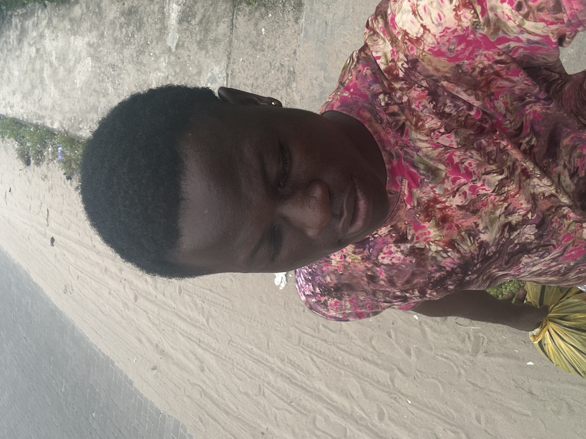 A dark-skinned young man stands on a sandy path next to a concrete sidewalk bordered by sparse vegetation in Badagry, Nigeria. He is wearing a short-sleeved t-shirt with a pink, purple, and brown tie-dye pattern. His left hand, visible in the lower left corner, clutches a yellow plastic bag. The man's head is tilted downwards, and his gaze is directed away from the camera, revealing a small gold stud earring in his right ear. The sandy path shows faint footprints and scattered debris, while the concrete sidewalk appears worn and weathered. The overall scene suggests an outdoor, casual setting, possibly near a beach or a less-developed area of the city.