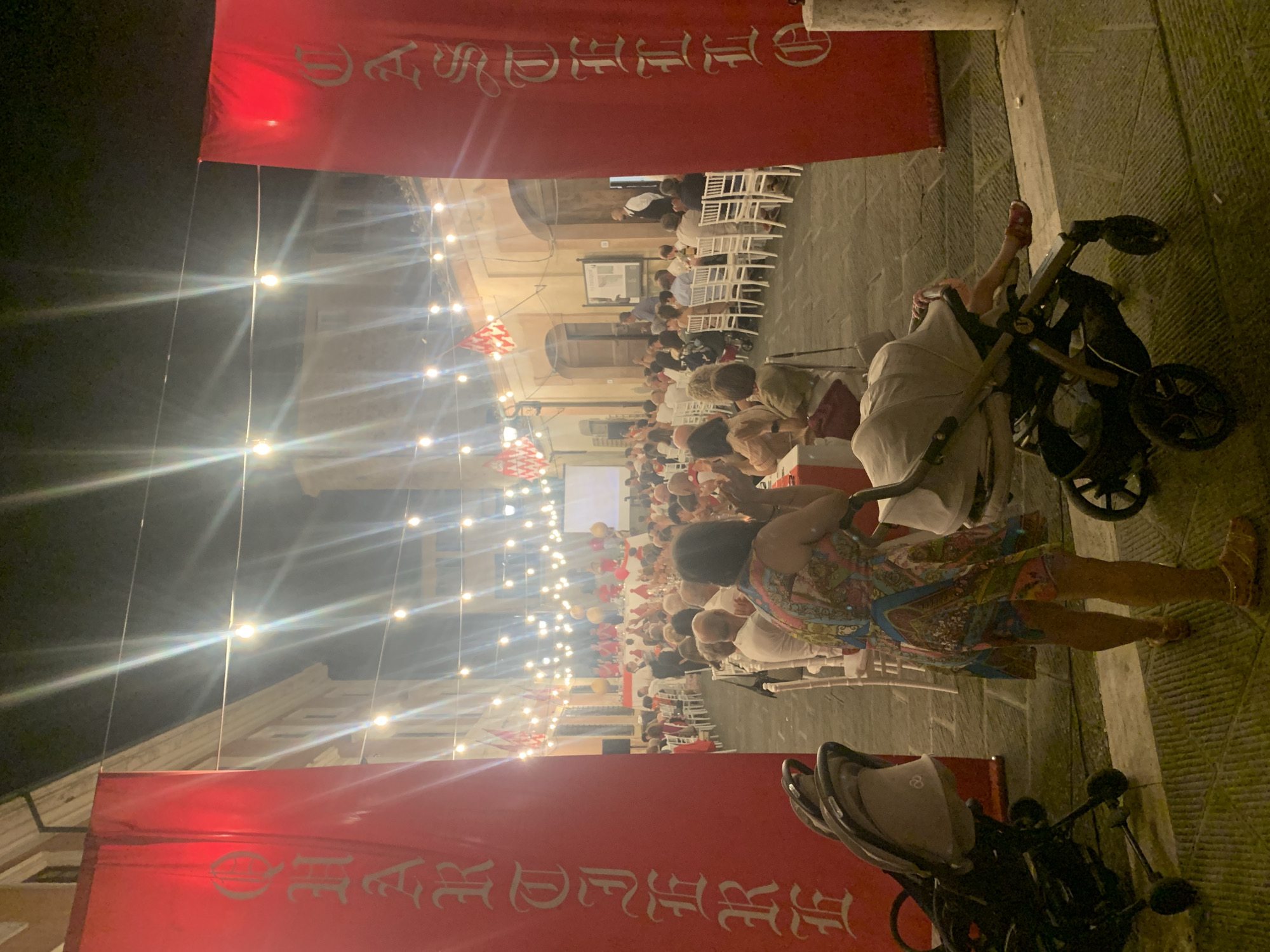 The image shows a nighttime outdoor event in San Quirico d'Orcia, Italy. In the foreground, two strollers are parked on a cobblestone street, with the legs and lower torso of a woman in a colorful dress visible between them. She is facing away from the camera and appears to be walking. In the background, a crowd of people is seated at tables, illuminated by string lights and red banners hanging from buildings. The banners on the left and right sides of the image have large, ornate white lettering. The overall atmosphere suggests a festive gathering or dinner event.