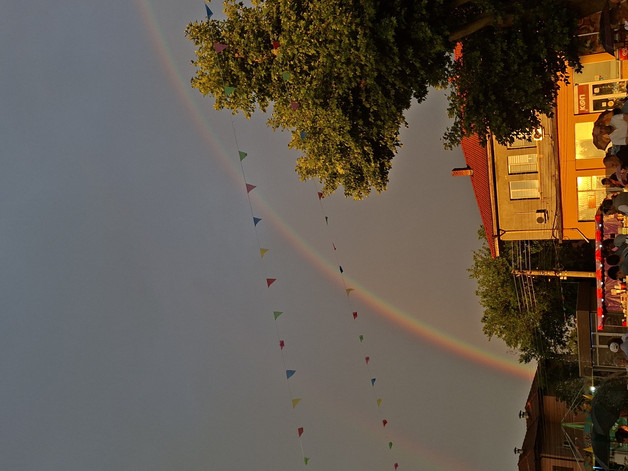 A low-angle, eye-level photograph depicts a street scene in the city of Yasen, Bulgaria, under an overcast sky featuring a prominent double rainbow. The time of day appears to be late afternoon or early evening, characterized by warm, orange light illuminating the lower parts of the scene, particularly the buildings.
The sky, occupying the majority of the upper frame, is a gradient of muted grey-blue at the top, transitioning to lighter, reddish-orange tones towards the horizon. A vibrant double rainbow arcs across the sky from left to right, with the primary bow being more distinct and colorful. Two parallel strings of multicolored triangular bunting flags (red, yellow, green, blue) are strung across the mid-ground, diagonally ascending from left to right, partially obscuring the rainbow.
On the right side of the image, a two-story building features a red-tiled roof and light ochre walls. Its ground floor is a commercial establishment, brightly lit with an intense orange glow emanating from its windows and entrance. A sign displaying Cyrillic letters "КОП" is visible. Two air conditioning units are affixed to the first-floor wall. A large, leafy green tree with illuminated foliage partially obscures the right side of this building and extends into the upper right corner.
On the far left, another building with a reddish-brown tiled roof and an awning or large umbrella structure is partially visible. Further back, another cluster of green trees is present.
In the foreground and mid-ground, numerous people are gathered on a paved area or street, primarily in front of the brightly lit commercial building on the right. They are standing, conversing, and moving about. Several individuals are beneath dark umbrellas, suggesting recent or ongoing light rain, or protection from the sun. A man in a white polo shirt is prominent in the lower-middle foreground, facing forward. Electrical utility poles and wires are visible on the right, traversing the space between the tree and the buildings. A street vendor's stall with a red awning and various items is present in the lower right corner, adjacent to the commercial building. A cooler or bin with a partial logo (possibly "ICE") is visible near the far right edge.
