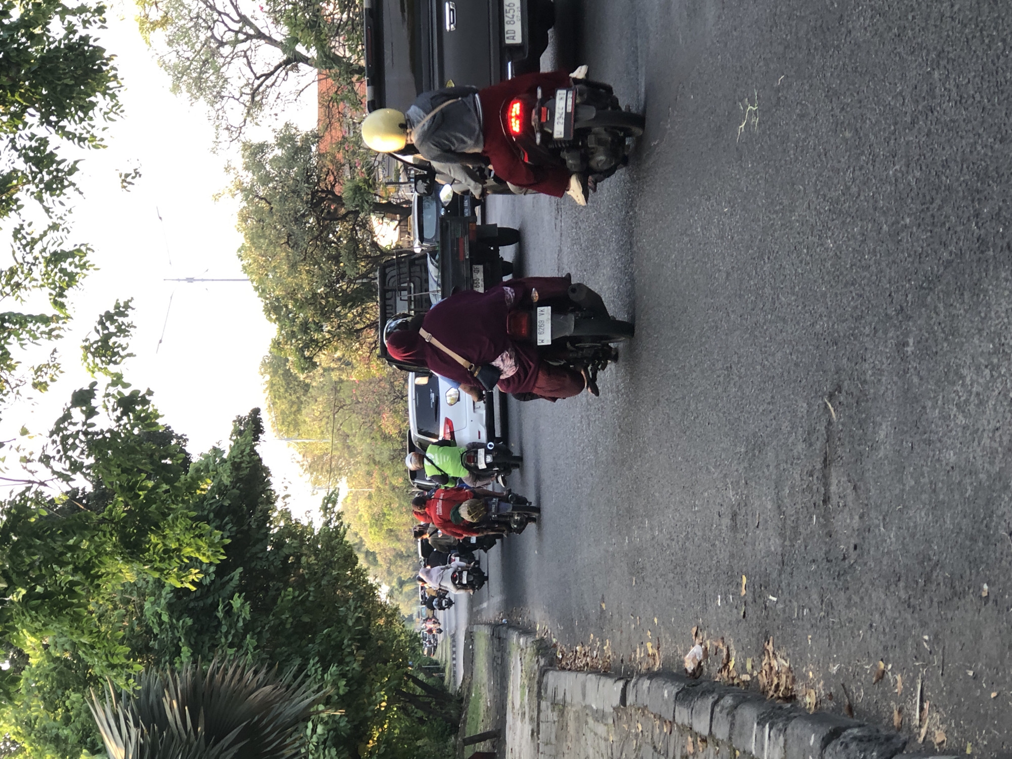 The image depicts a street scene in Sumorame, Indonesia, likely during the late afternoon due to the warm, low-angle sunlight illuminating the trees and road. A line of motorcycles and a few cars are moving along a paved road. Several people are riding on the motorcycles, most wearing helmets.
In the foreground, two motorcycles are prominently featured. The motorcycle on the right is ridden by a person wearing a yellow helmet and a gray jacket, with dark red pants. The motorcycle on the left is ridden by a person wearing a dark colored helmet and a maroon headscarf with a matching maroon robe. Both motorcycles have visible license plates.
Further down the road, other motorcycles and a white car are visible, indicating moderate traffic. Trees line the left side of the road, casting shadows and creating a natural canopy. The road has a distinct curb on the left side, with some dry leaves and debris scattered on it. The overall environment suggests a typical urban or suburban street in Indonesia with active daily life and transportation.