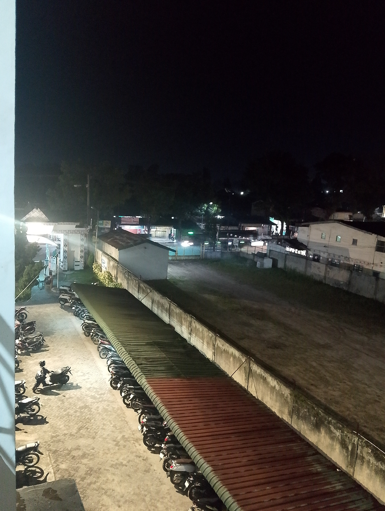 The image depicts an outdoor scene at night in the city of Pematang Siantar, Indonesia, with a focus on a parking area filled with motorcycles. The parking lot is paved with interlocking concrete blocks and is partially covered by a long, corrugated metal roof, predominantly red and green, providing shelter. A concrete wall runs along the edge of a grassy area adjacent to the parking lot.
On the left side of the image, a cluster of motorcycles is parked. Several are dark in color, with visible handlebars and wheels. One motorcycle in the foreground, on the far left, is being pushed by a person wearing dark clothing and a helmet.
Further back, under a bright white canopy structure with ornate white pillars and decorative elements, what appears to be a gas station or a similar service area is visible. Lights illuminate this area, casting reflections and creating a bright contrast against the night sky. Another building, a light-colored structure with a sloped roof, is situated behind the gas station.
In the background, a line of buildings can be seen, illuminated by lights, suggesting a more developed urban area. Trees and foliage are scattered throughout the scene, some silhouetted against the dark sky. The overall atmosphere is one of a typical urban night scene, with the primary activity revolving around the parked motorcycles and the illuminated service area.