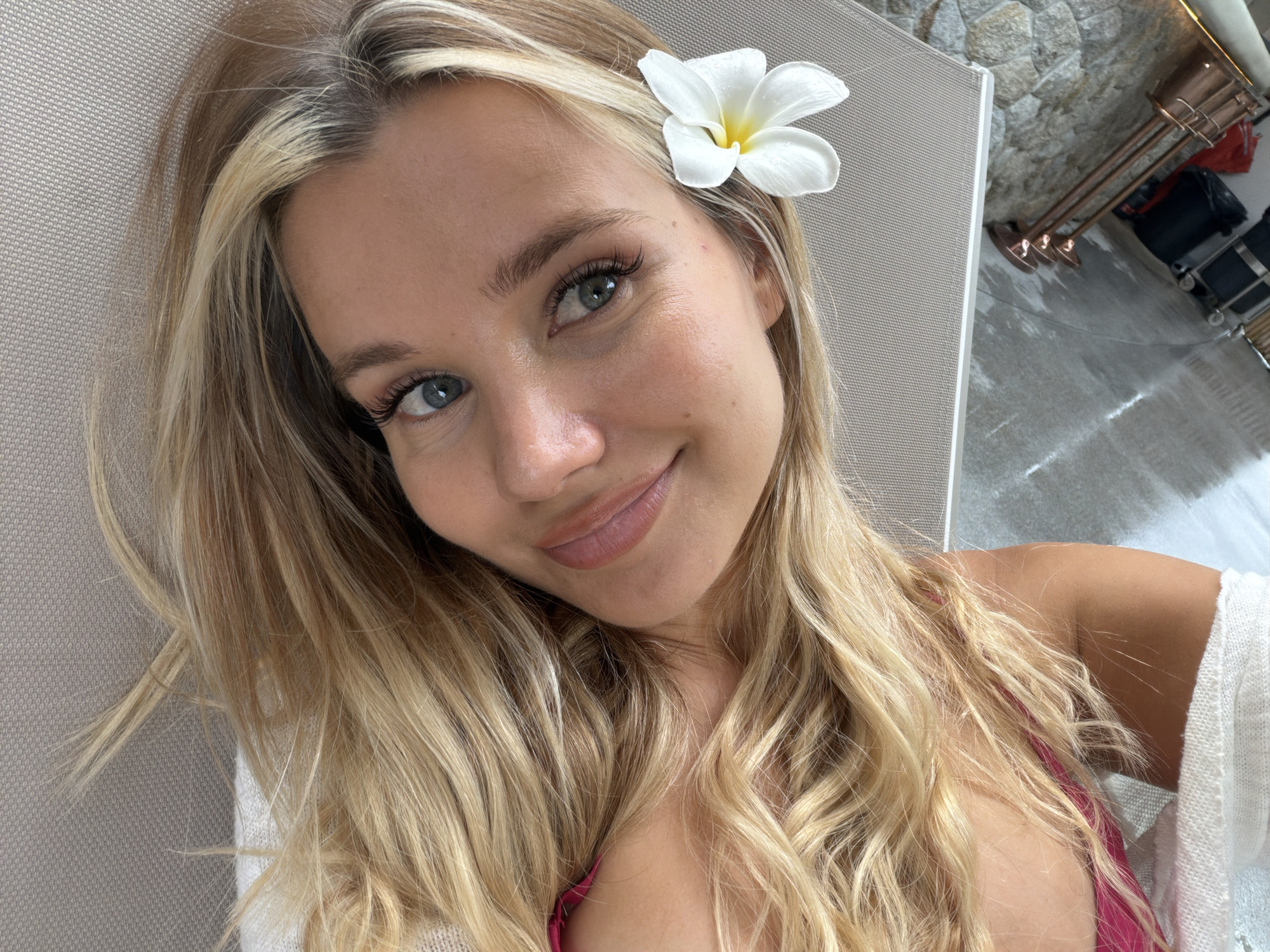 A blonde woman with blue eyes and light skin is lying on a textured surface, her head tilted towards the camera. She is wearing a pink top. A white flower with a yellow center is tucked behind her right ear. Her blonde hair is long and wavy, spread out around her head. The background shows a textured gray surface, a glimpse of stone wall, and a metal fixture with three lights. The overall scene suggests an outdoor or spa-like environment, potentially in Choeng Thale, Thailand, given the presence of tropical flora.