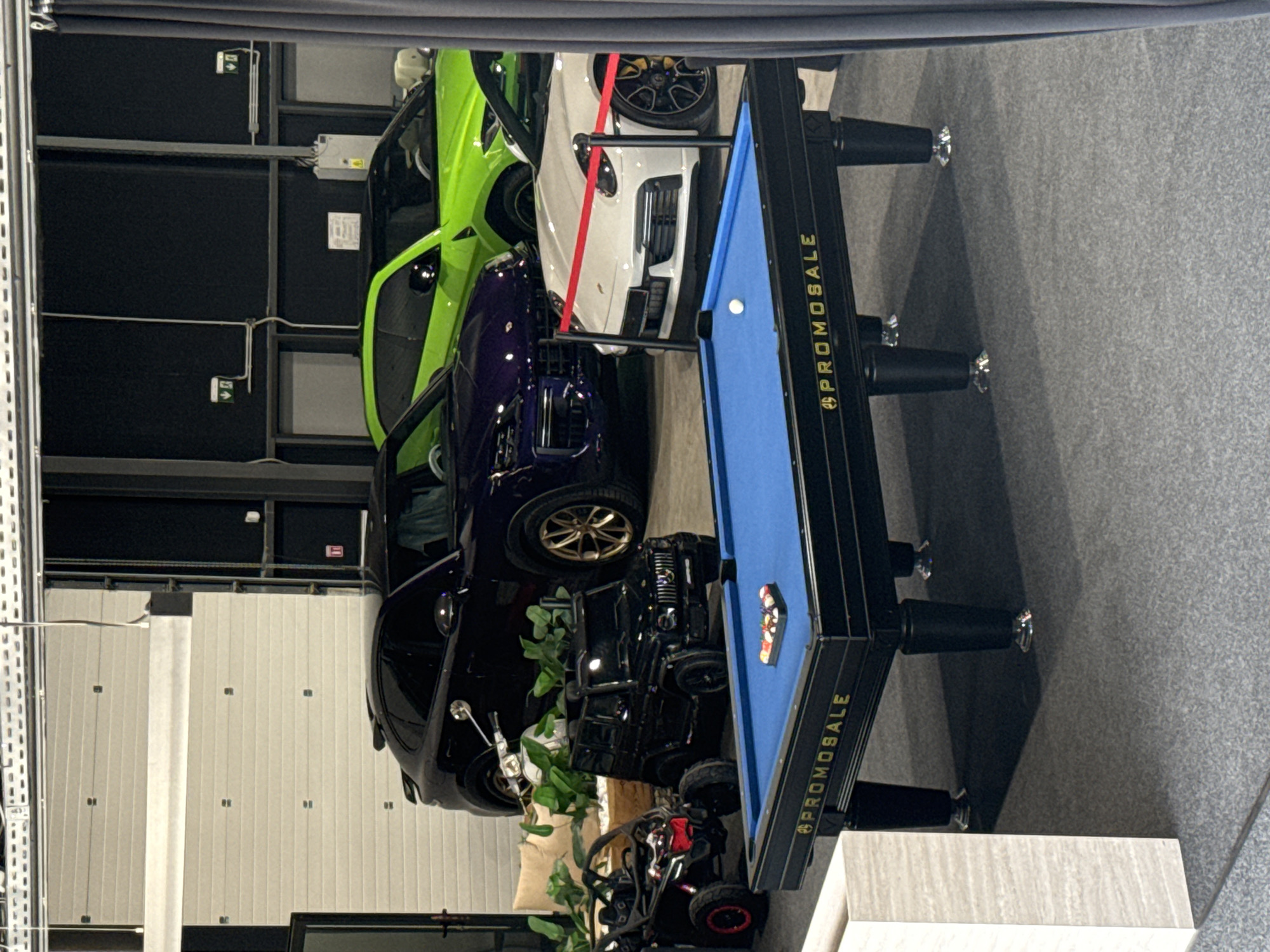 The image displays an indoor scene, likely a high-end garage, car showroom, or private collection space. Several luxury vehicles are prominently featured, arranged alongside recreational items.
In the foreground, from left to right (assuming the image is rotated 90 degrees clockwise for correct orientation), there is a bright lime green SUV, resembling a Lamborghini Urus. Next to it is a white sports car, possibly a Porsche, with a red strap visible over its hood. A dark purple SUV, similar to a Rolls-Royce Cullinan, is parked behind the white car. Further back, a black SUV, reminiscent of a Mercedes-Benz G-Wagen, is visible, with golden-colored wheel rims. A black car, possibly another SUV or a sedan, is partially seen behind the black G-Wagen.
A blue-felt billiards table with a black frame stands in the middle ground. A white cue ball is visible on the playing surface, and a rack of colored billiard balls is neatly placed in one of the pockets. The word "PROMOSALE" is clearly visible in gold lettering along the side of the table frame.
Other objects in the scene include a small black ATV or buggy and a white scooter or mini-motorcycle situated near the black SUV. Green leafy plants in pots are interspersed among the vehicles, adding a touch of greenery. The background consists of tall, segmented white garage doors and dark walls with visible electrical conduits and emergency exit signs. The floor is covered in a dark grey, textured material. No people are present, and no specific activity is taking place, suggesting the scene is set up for display or storage. The lighting is artificial, making the time of day indeterminate.