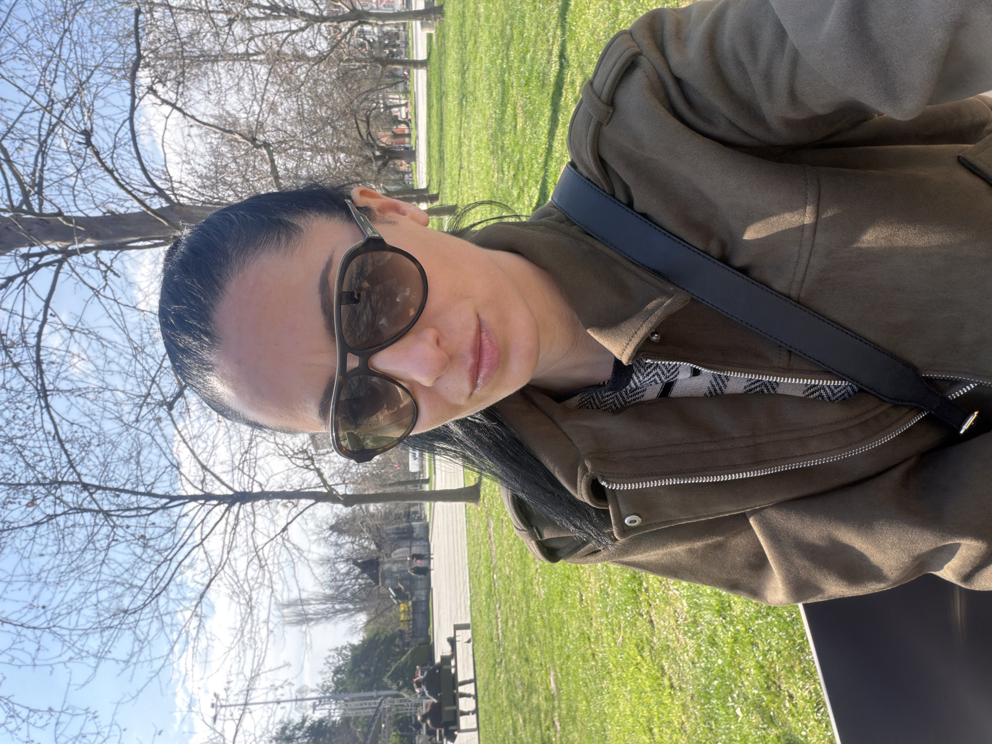 The image shows a woman wearing sunglasses and a jacket, outdoors in a park-like setting. The background features trees, grass, and a building in the distance. Given the location specified as Pleven, Bulgaria, and the landmarks visible in the background, it is likely the scene is in the Kaylaka Park.