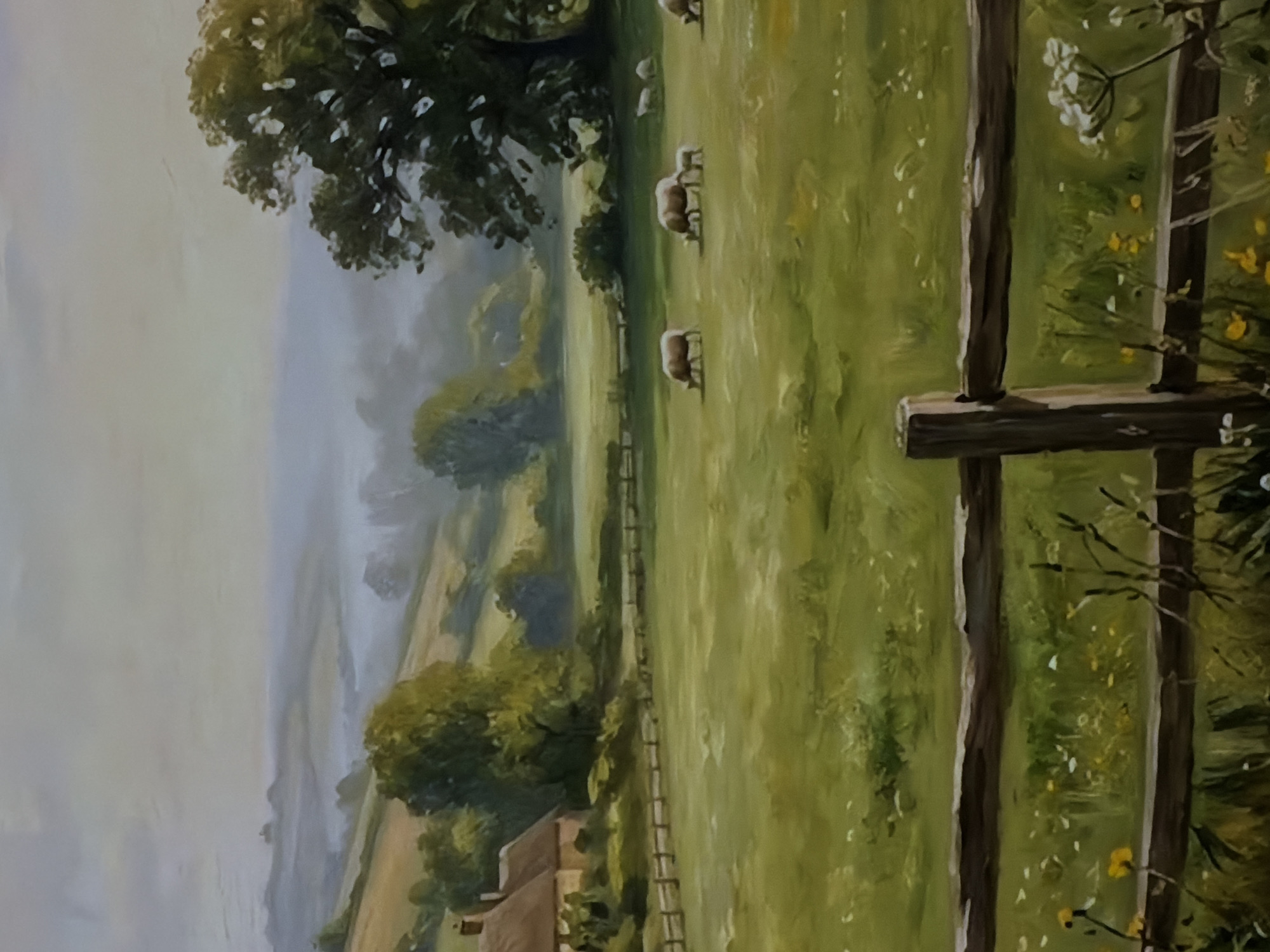 This is a landscape painting, depicted rotated 90 degrees counter-clockwise in the provided image. When viewed correctly, it portrays a serene pastoral scene.
**Subjects:** The primary subjects are a flock of sheep, trees, green fields, a wooden fence, and a distant house.
**Scene and Setting:** The setting is a rolling countryside, possibly in the early morning or late afternoon given the soft, diffused light and hazy distant hills.
**Foreground:** On the right (when viewed correctly), a rustic wooden fence stretches across the scene, with close-up details of its posts and horizontal rails. Sprigs of white and yellow wildflowers, along with other green foliage, are visible growing near the fence, suggesting a vibrant, natural environment. The immediate foreground consists of lush green grass.
**Midground:** The central part of the painting features a large, verdant field where several sheep are grazing peacefully. There are at least three prominent sheep, with others appearing smaller and further away in the field. Another fence line, less distinct, runs horizontally across the midground, separating different sections of the landscape.
**Background:** In the distance, rolling hills and valleys covered with dense trees and fields fade into a soft, atmospheric haze, creating a sense of depth and tranquility. A small house with a reddish-brown roof is nestled among the trees in the lower-left background.
**Activity or event:** The main activity is sheep grazing in the field, contributing to the overall sense of peace and rural life.
**Notable details:**
* **Time of day/weather:** The soft lighting and distant haze suggest either dawn or dusk, or a slightly overcast day, creating a gentle and calm atmosphere.
* **Location cues:** The landscape resembles a traditional European countryside scene, likely a temperate climate. There are no specific visual cues within the painting to indicate the location as Sofiivska Borshchahivka, Ukraine, or any other specific geographical region.
* **Artistic Style:** The painting exhibits a realistic or impressionistic style, with visible brushstrokes, particularly in the foliage and grass, adding texture and life to the scene.
**Visible text:** There is no visible text in the image.
